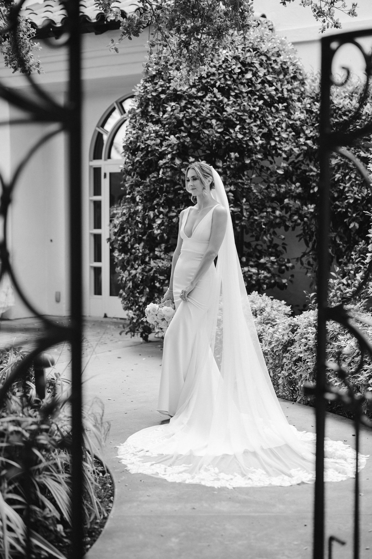 Black and white photo of wedding dress with cathedral train in garden setting framed by iron gates.
