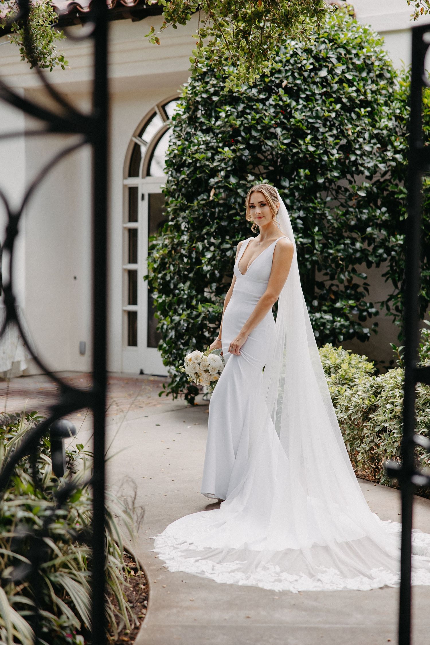 Stunning bridal portrait in sleek white gown against lush garden backdrop with arched doorway.
