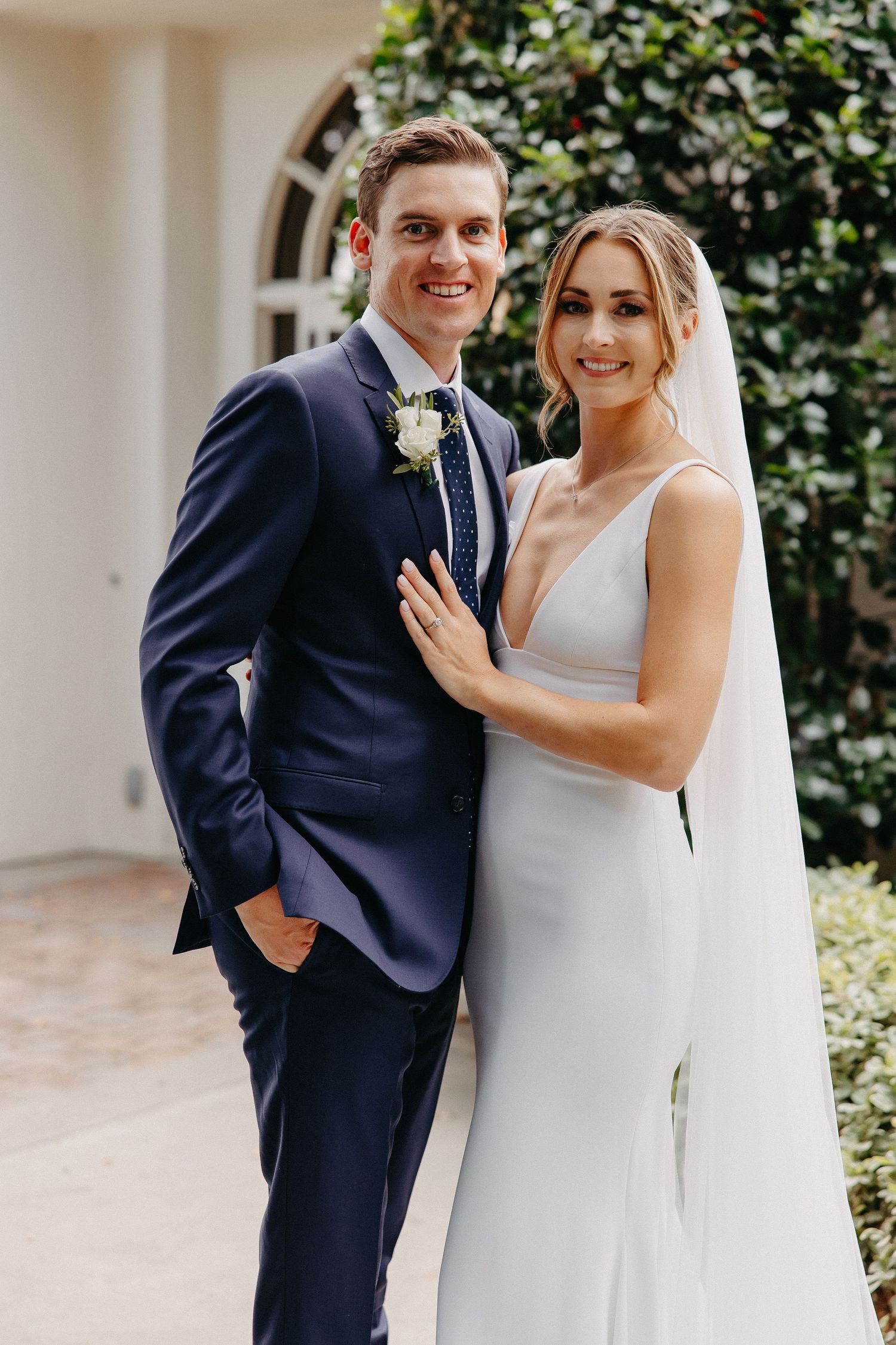 Wedding couple poses in garden, wearing navy suit and white gown with cathedral veil.
