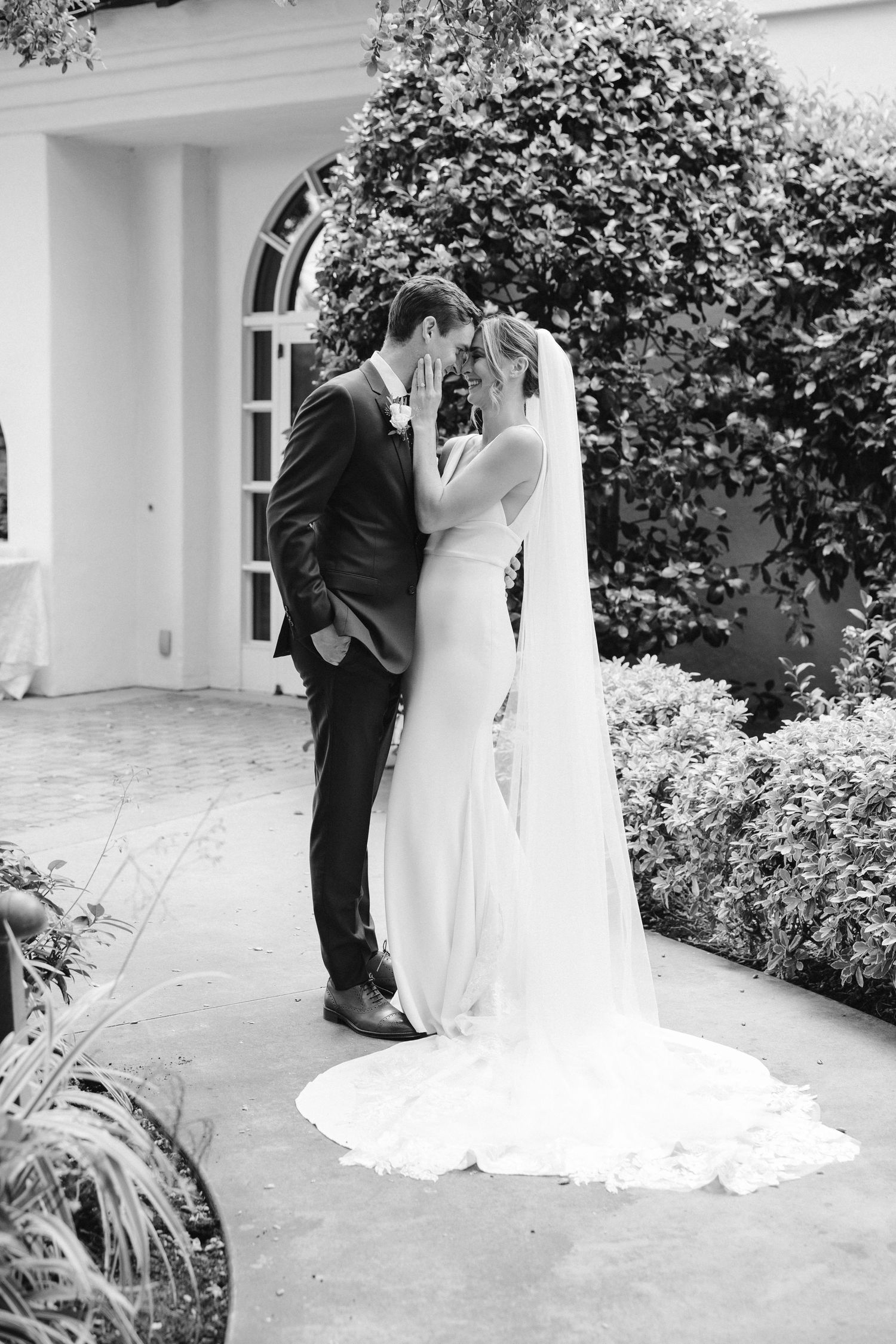 Black and white romantic wedding portrait in front of arched doorway and greenery.