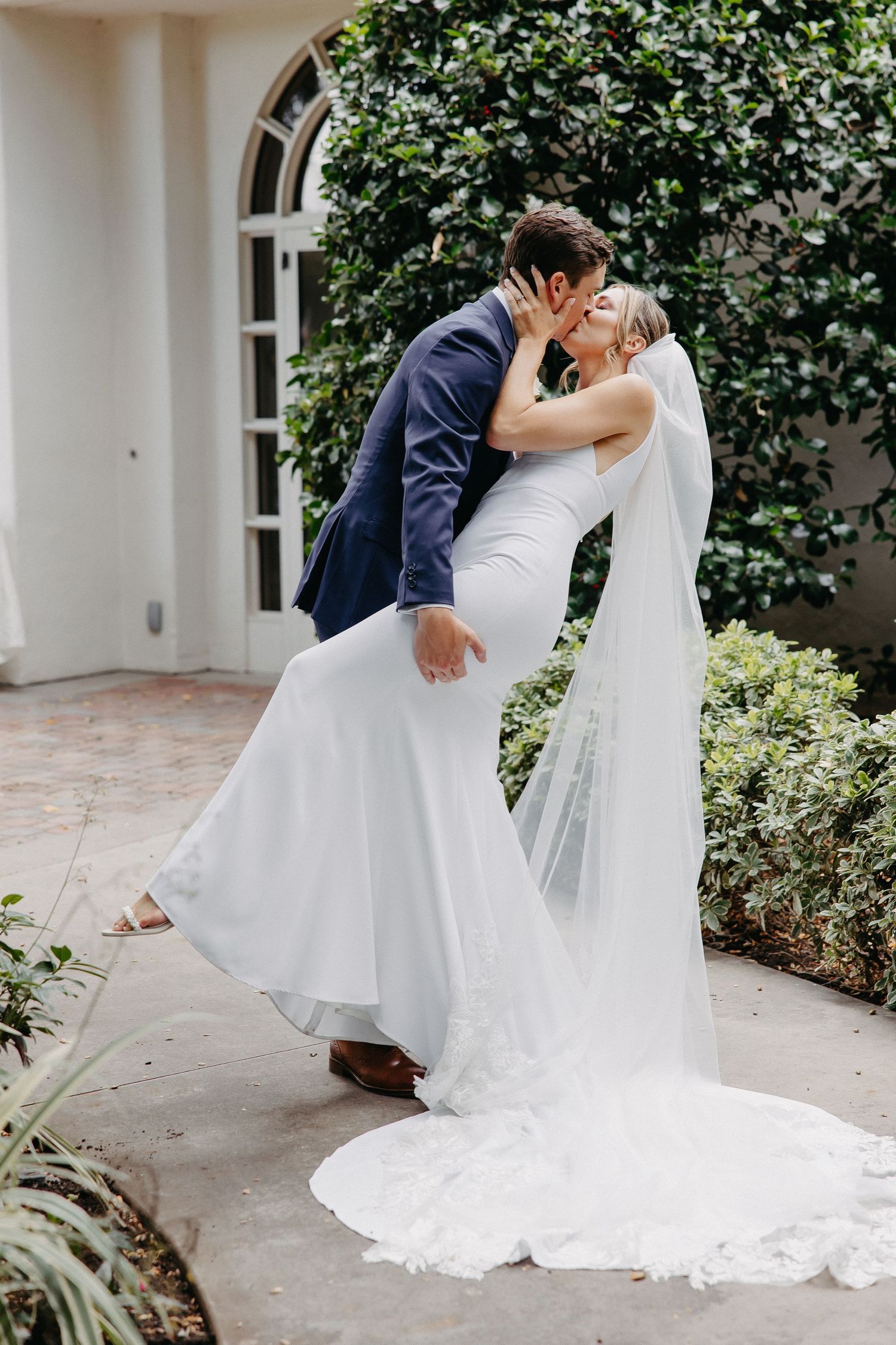 Romantic dip kiss wedding photo with flowing veil against garden backdrop.