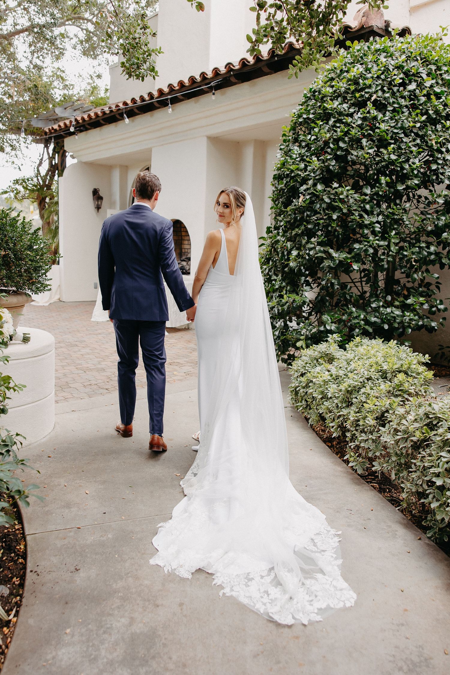 A couple walking down a garden path with a flowing white wedding dress trailing behind.
