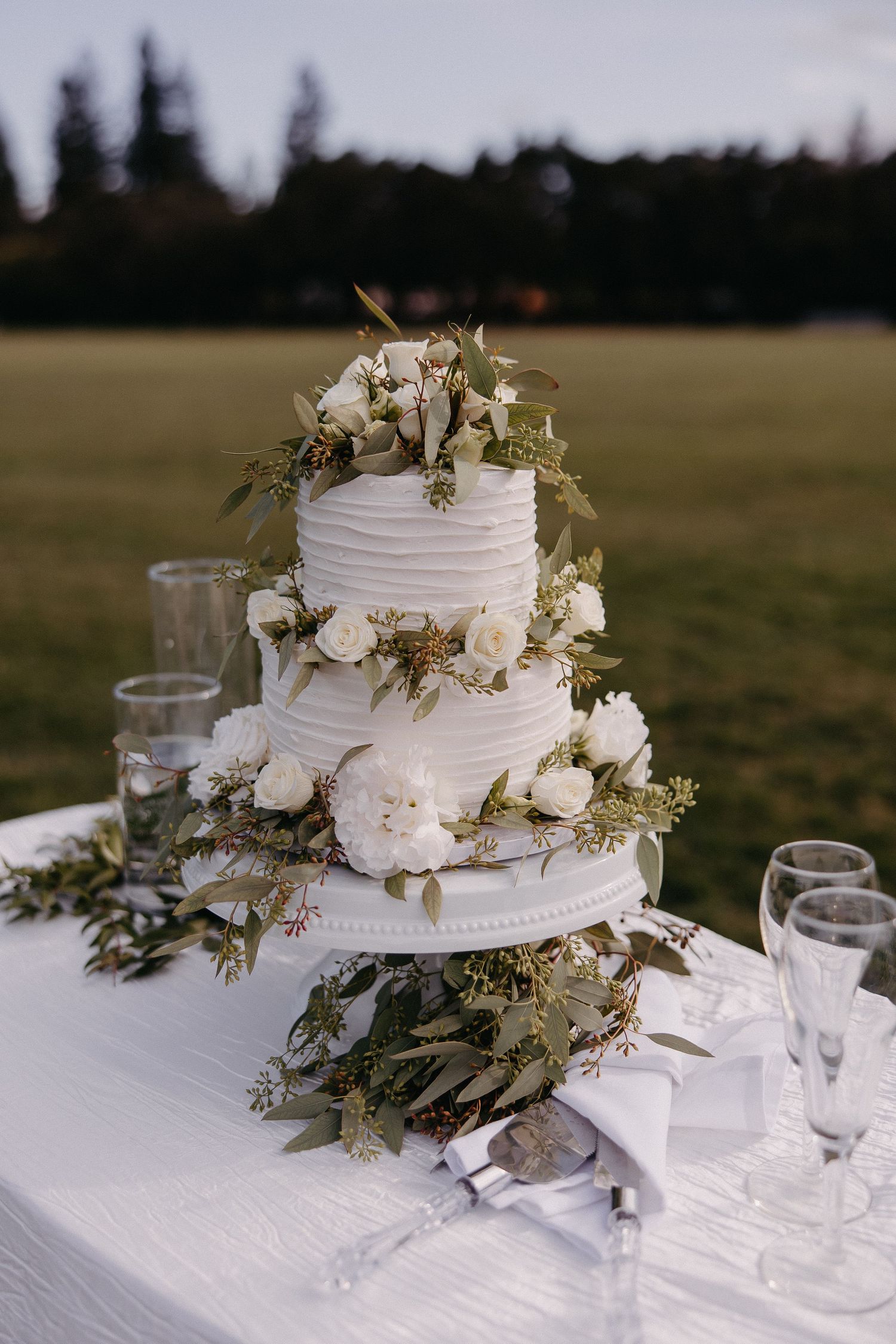 Elegant three-tier white wedding cake decorated with fresh flowers and greenery on an outdoor table.