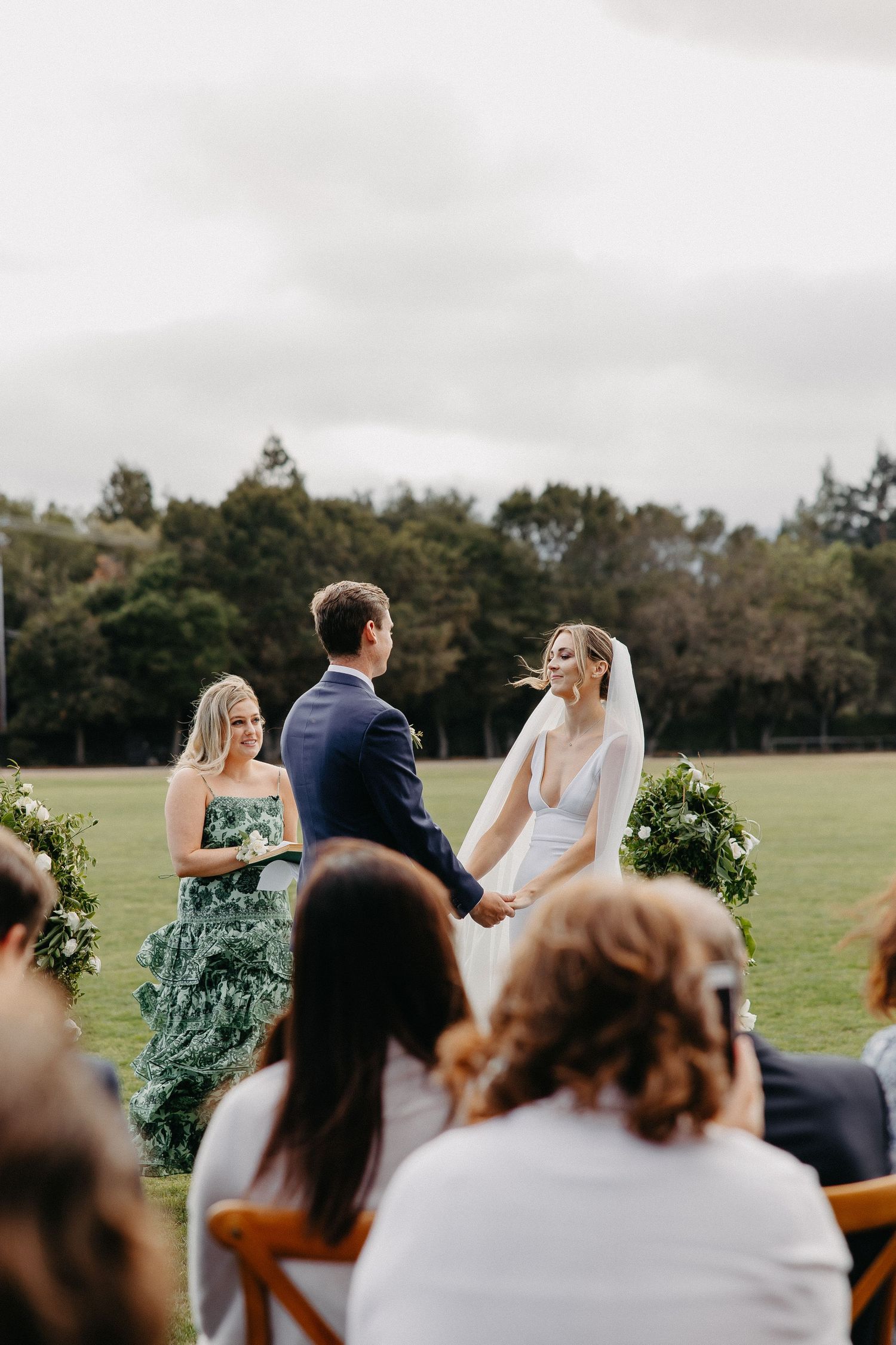 Wedding ceremony in progress on a green lawn with floral columns and seated guests.