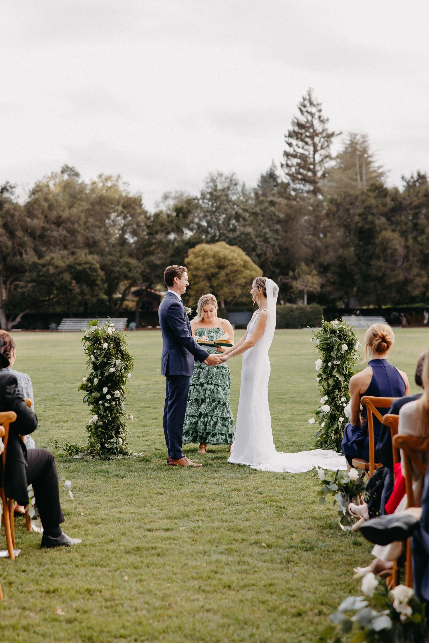 Outdoor wedding ceremony with floral columns and wooden chairs on a grassy field.