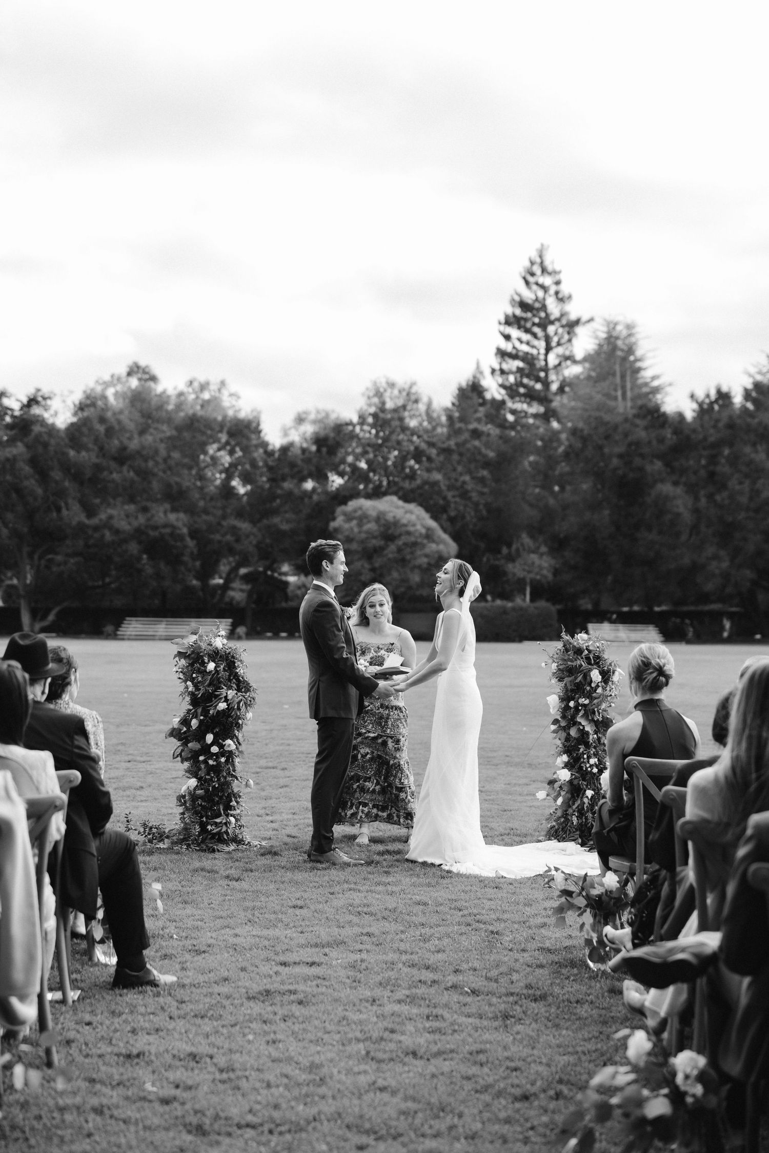 Black and white photo of wedding ceremony with guests seated on either side of flower-lined aisle.