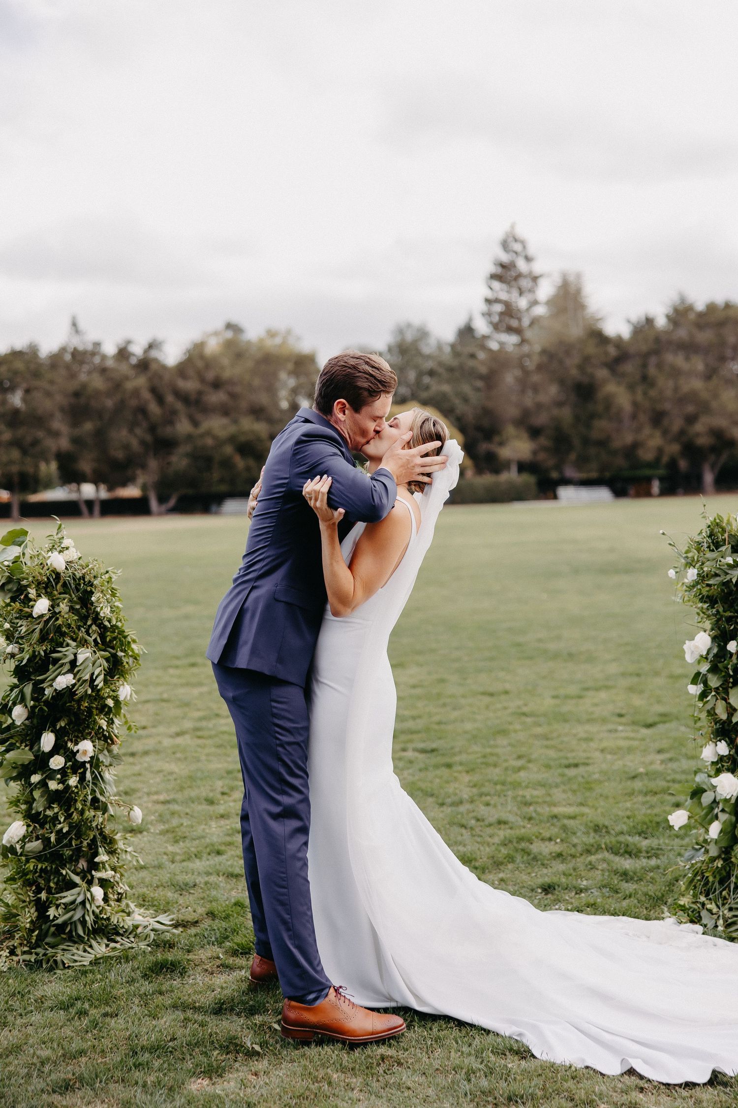 Romantic first kiss between newlyweds at outdoor garden wedding with floral arch.