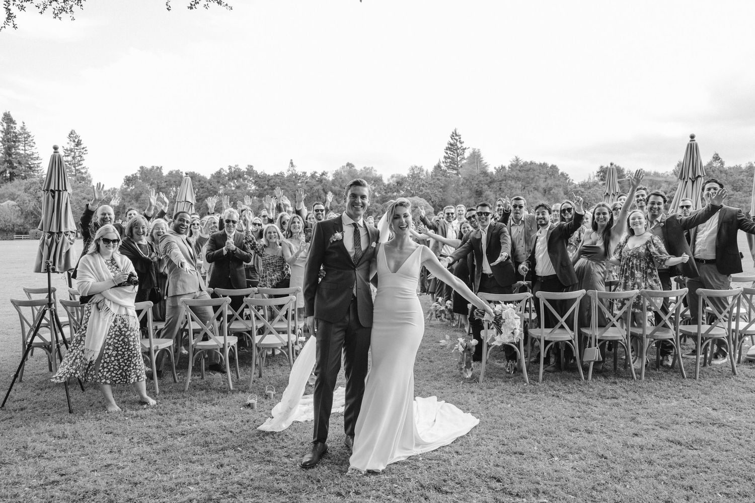 Black and white photo of wedding ceremony with guests seated on wooden chairs outdoors.