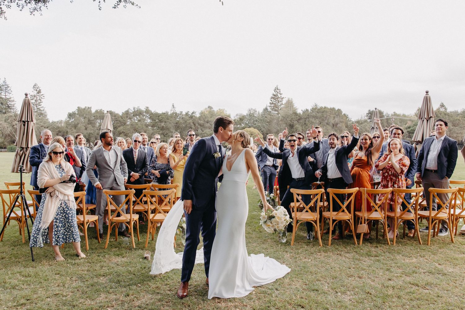 Outdoor wedding ceremony with wooden chairs and standing guests celebrating the couple.
