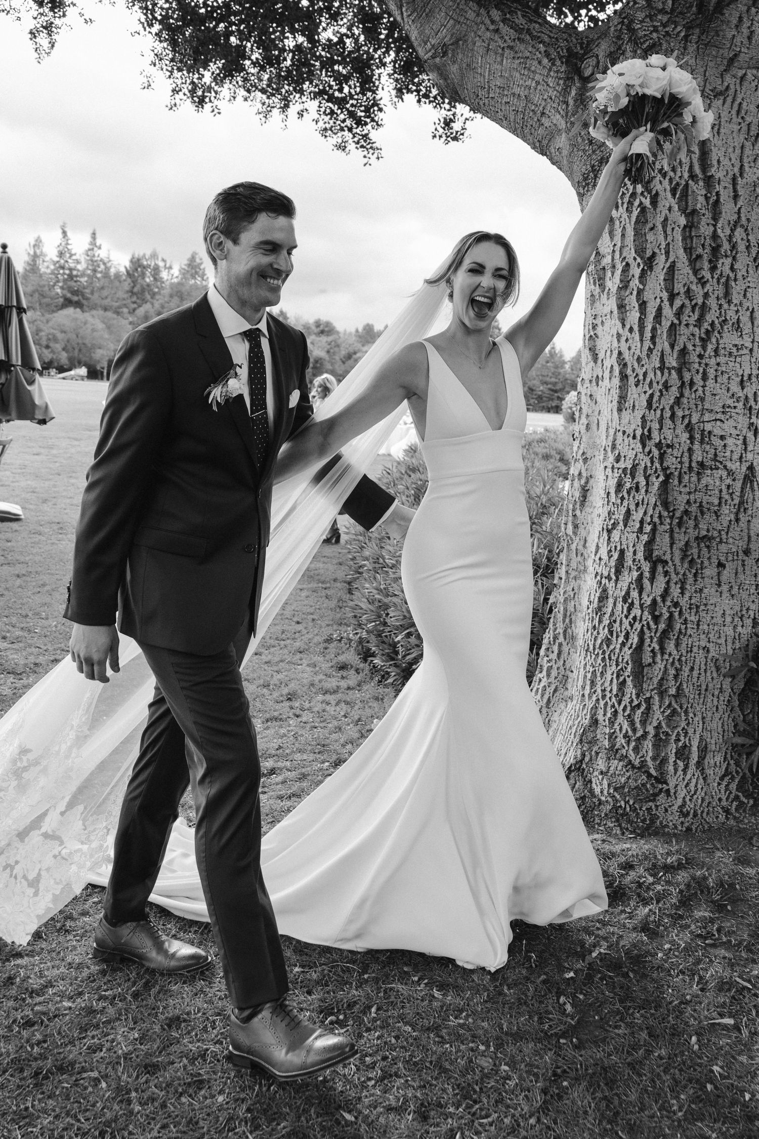 Black and white candid of newlyweds celebrating by large tree with flowing veil.