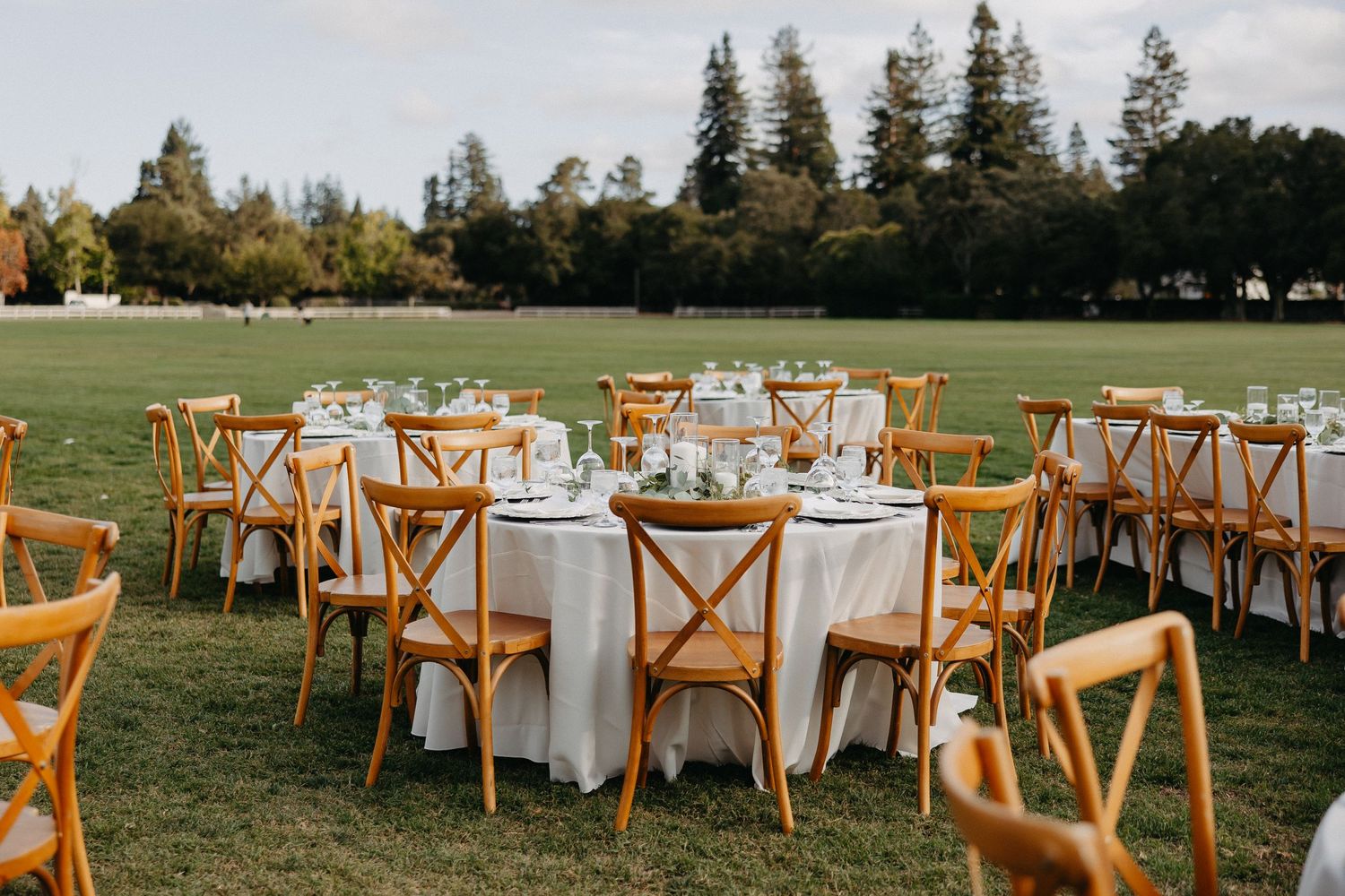 Rustic outdoor wedding reception setup with wooden crossback chairs and white linens.