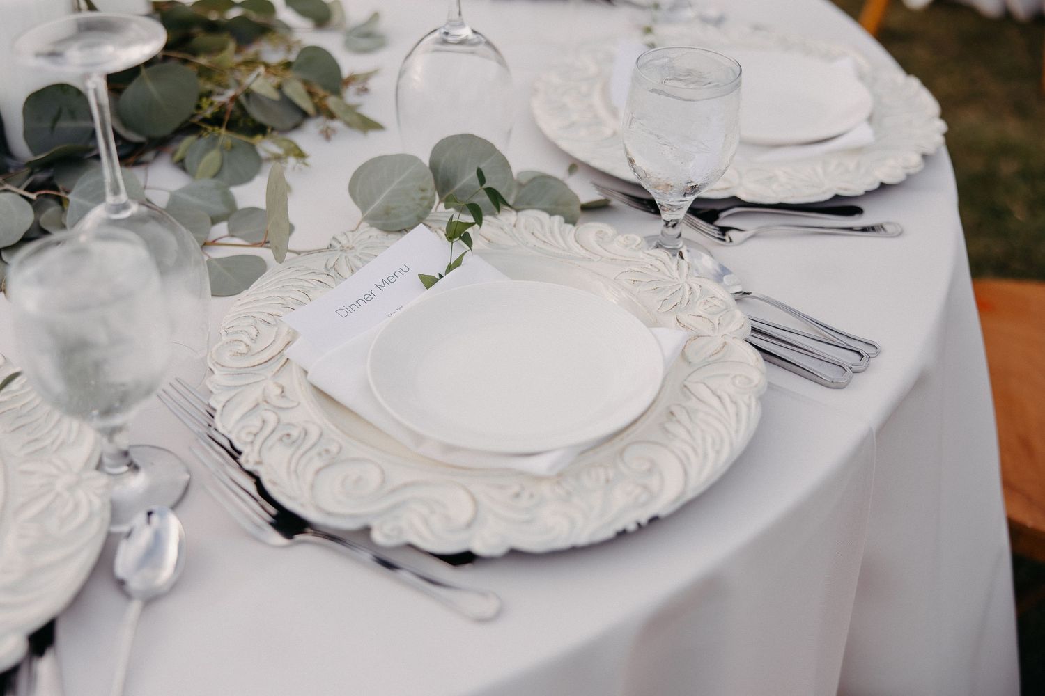 Elegant table setting with ornate white charger plate, eucalyptus leaves, and formal silverware on white tablecloth.