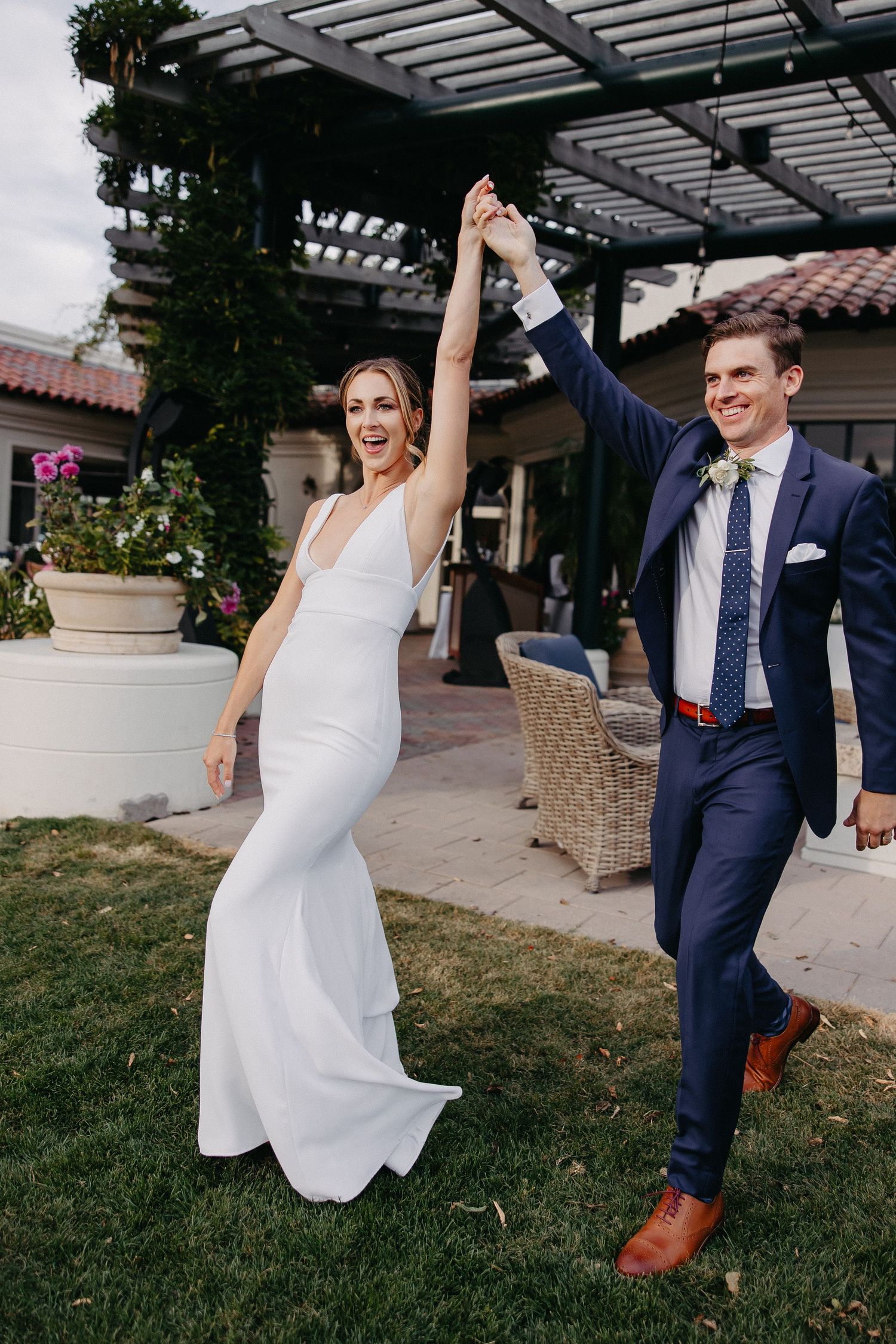 Newlyweds celebrating with raised hands as they walk on grass near a covered patio.