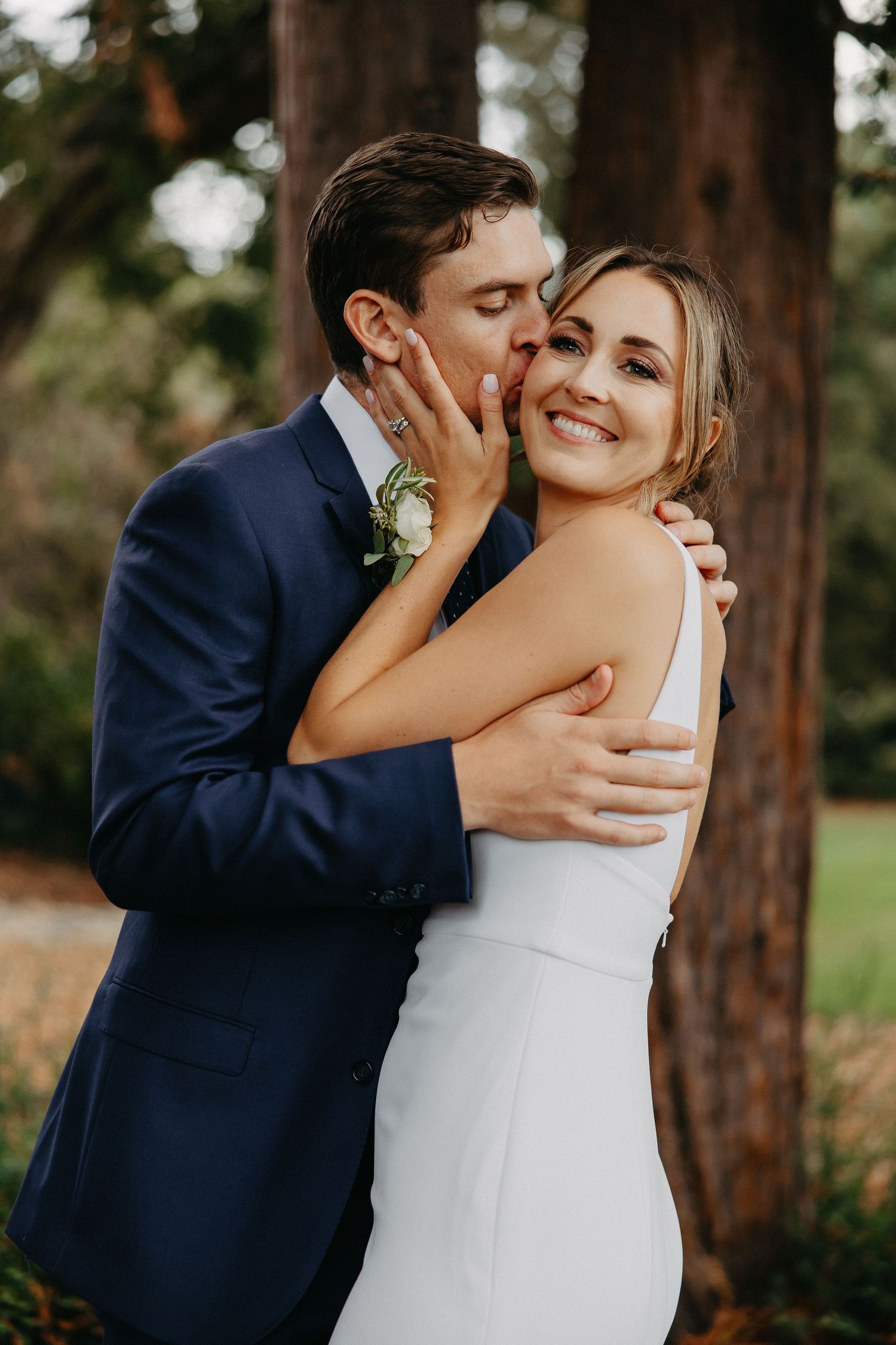 Sweet candid moment between couple in wedding attire surrounded by tall trees.