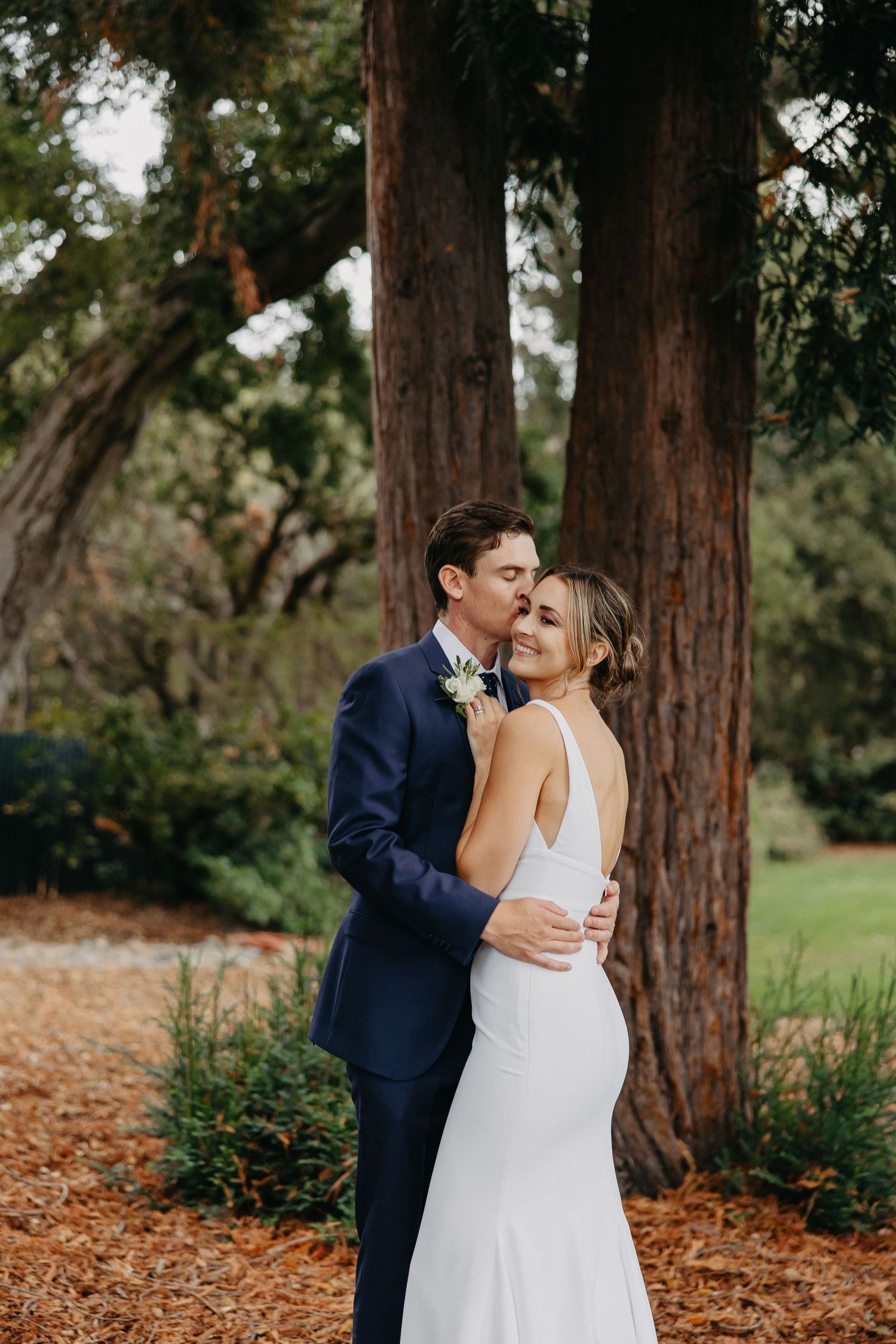 Elegant couple embraces on rustic woodland path with tall trees in background.
