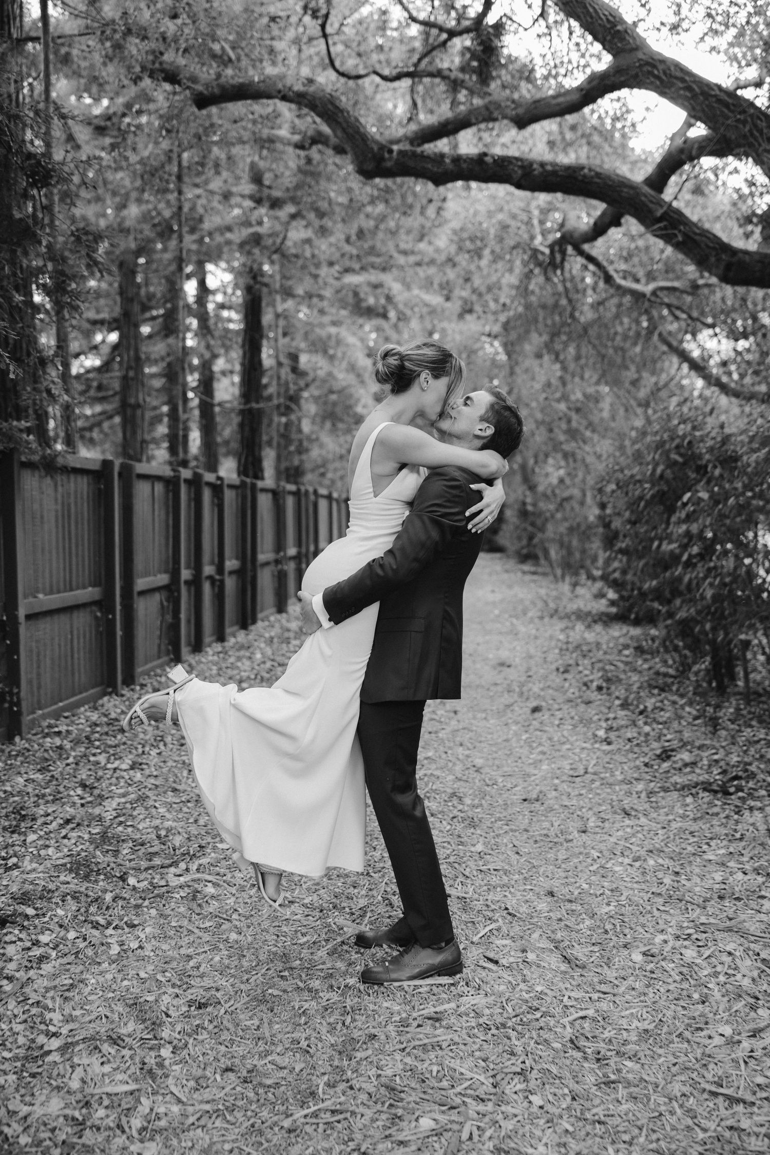 Black and white photo of romantic lift pose on tree-lined path with fence.