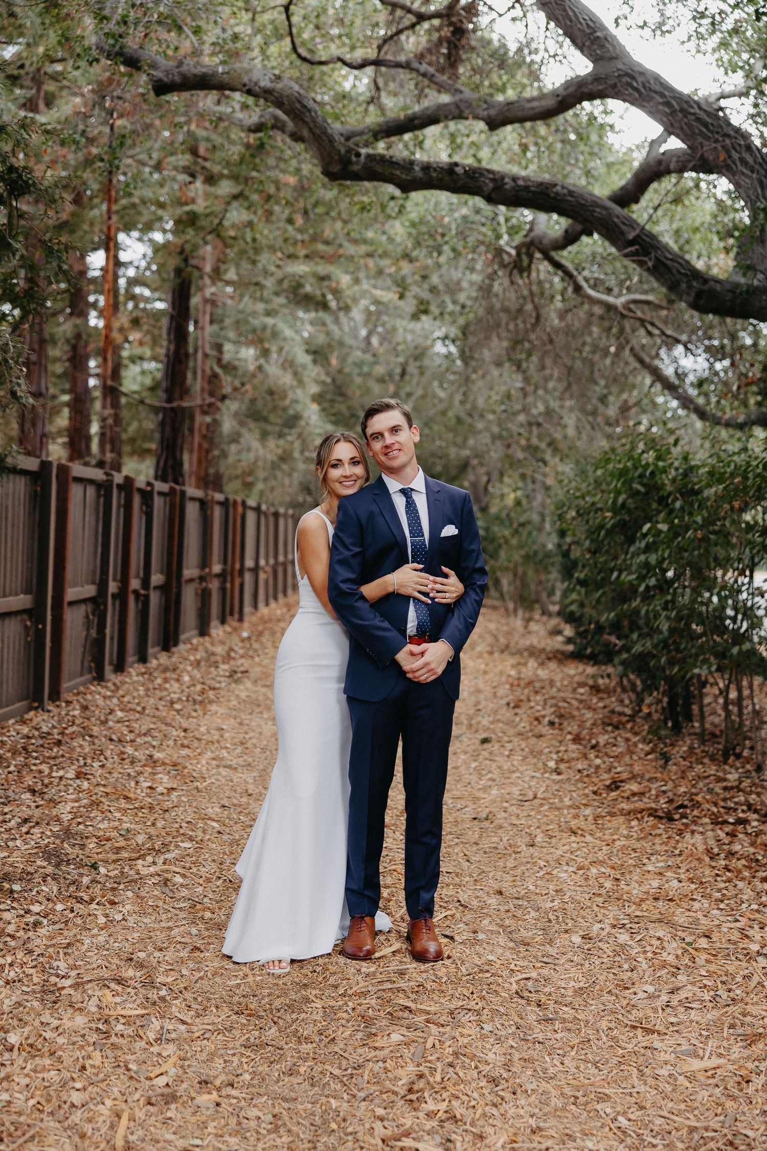 Stylish wedding couple poses on rustic path surrounded by majestic trees and iron fence.