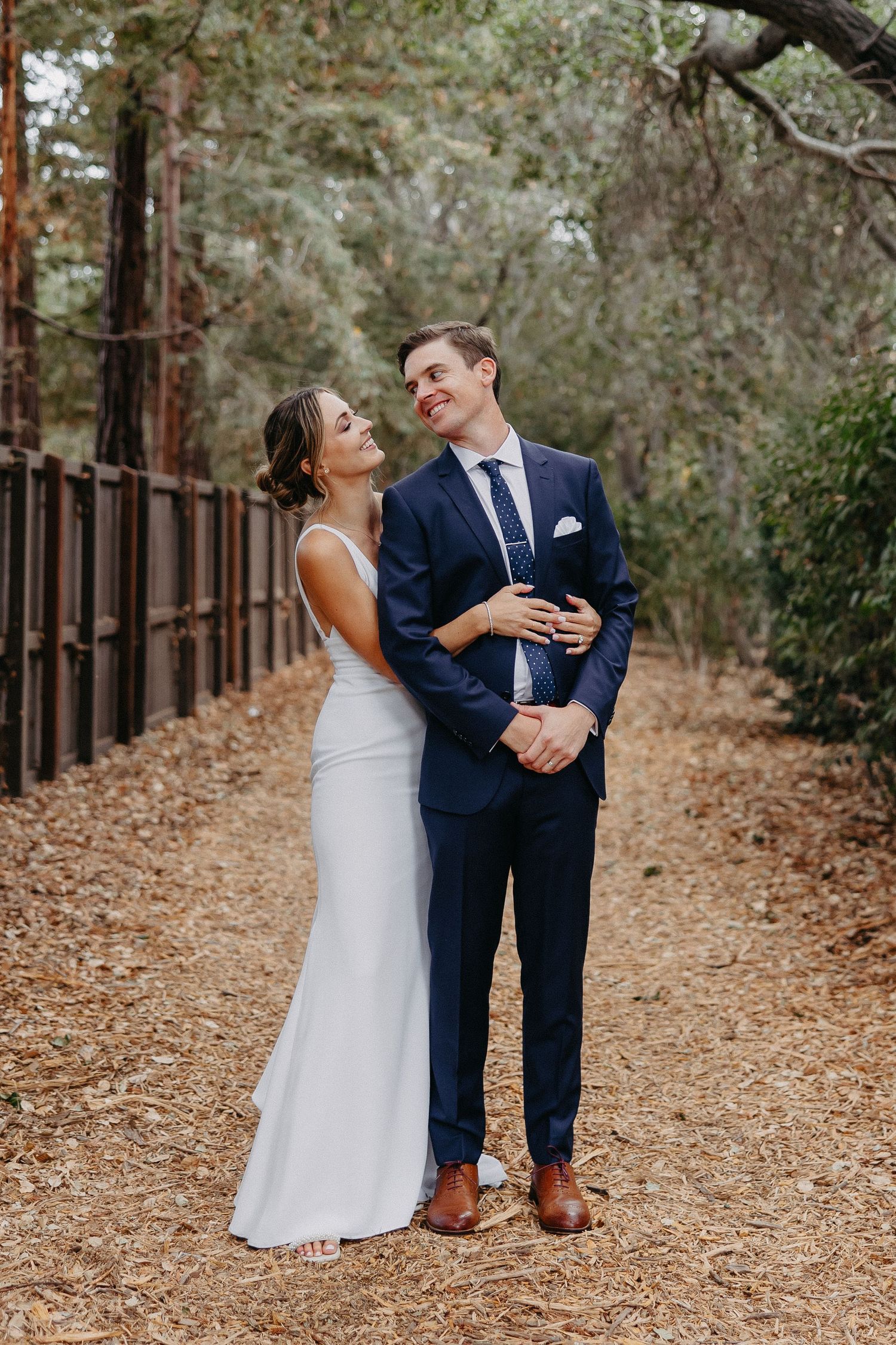 Happy couple shares loving moment on leaf-covered forest path with iron fence backdrop.