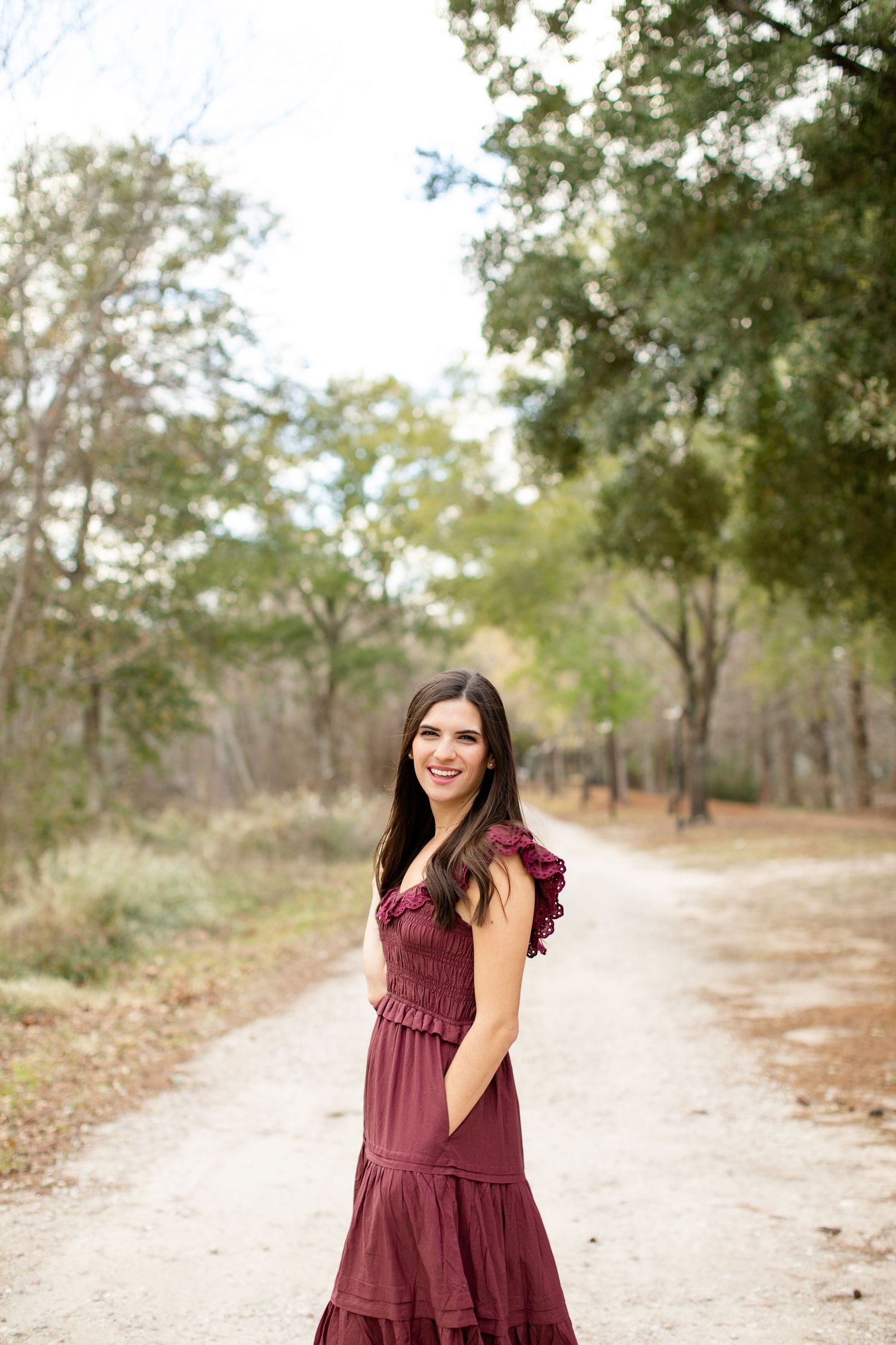 Woman in flowing burgundy dress stands on dirt path through wooded area.