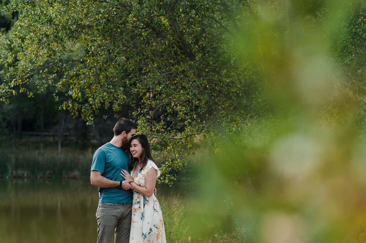 A couple shares an intimate moment by a tranquil pond with sunlit foliage in the background.