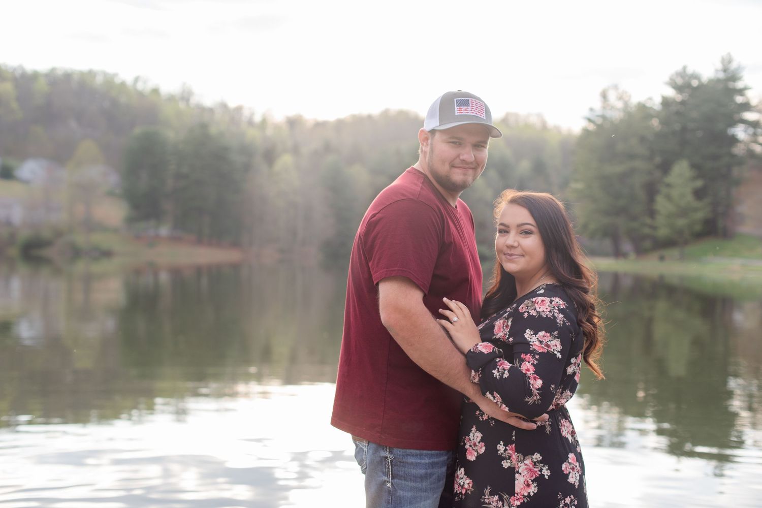 Happy couple embraces while standing on wooden dock with serene lake and forest backdrop.