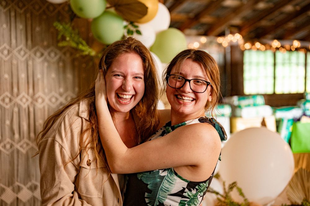 Two women celebrating at a festive party with green balloons and decorative backdrop.