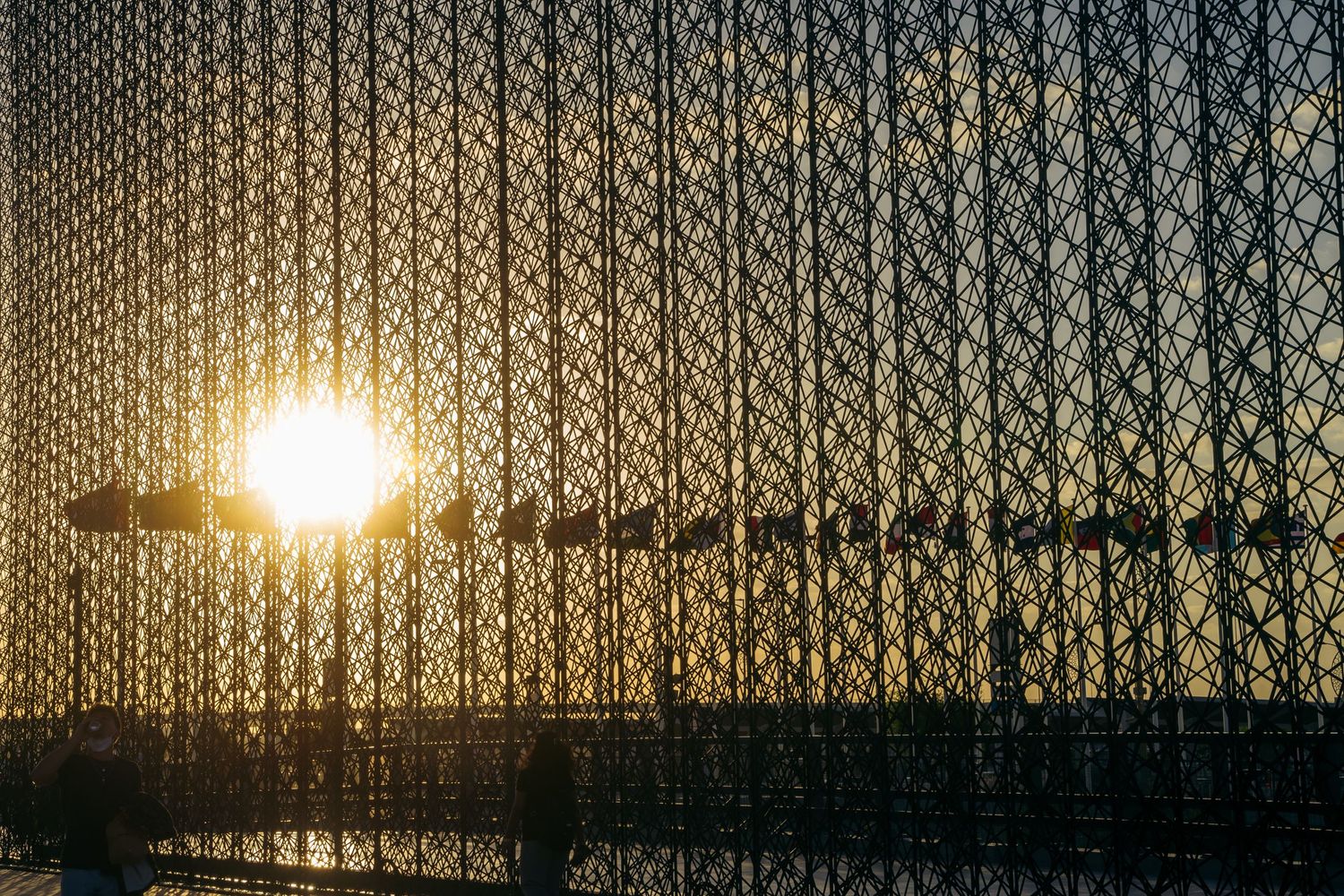 Sun setting behind geometric lattice fence structure creating dramatic silhouette and golden light.