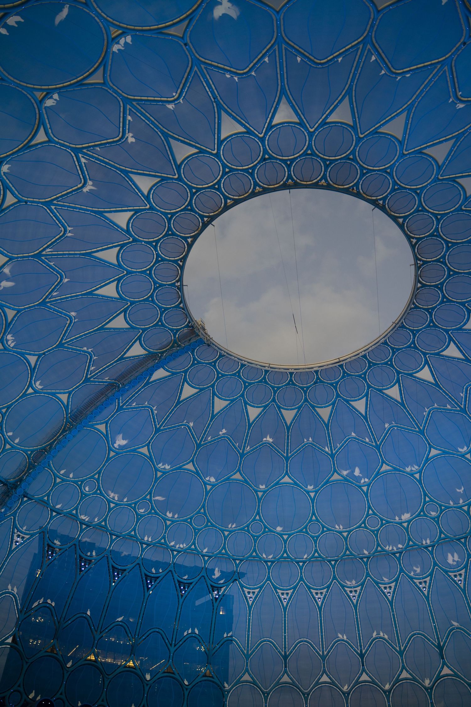 Upward view of a blue geometric dome ceiling with circular skylight and intricate petal patterns.
