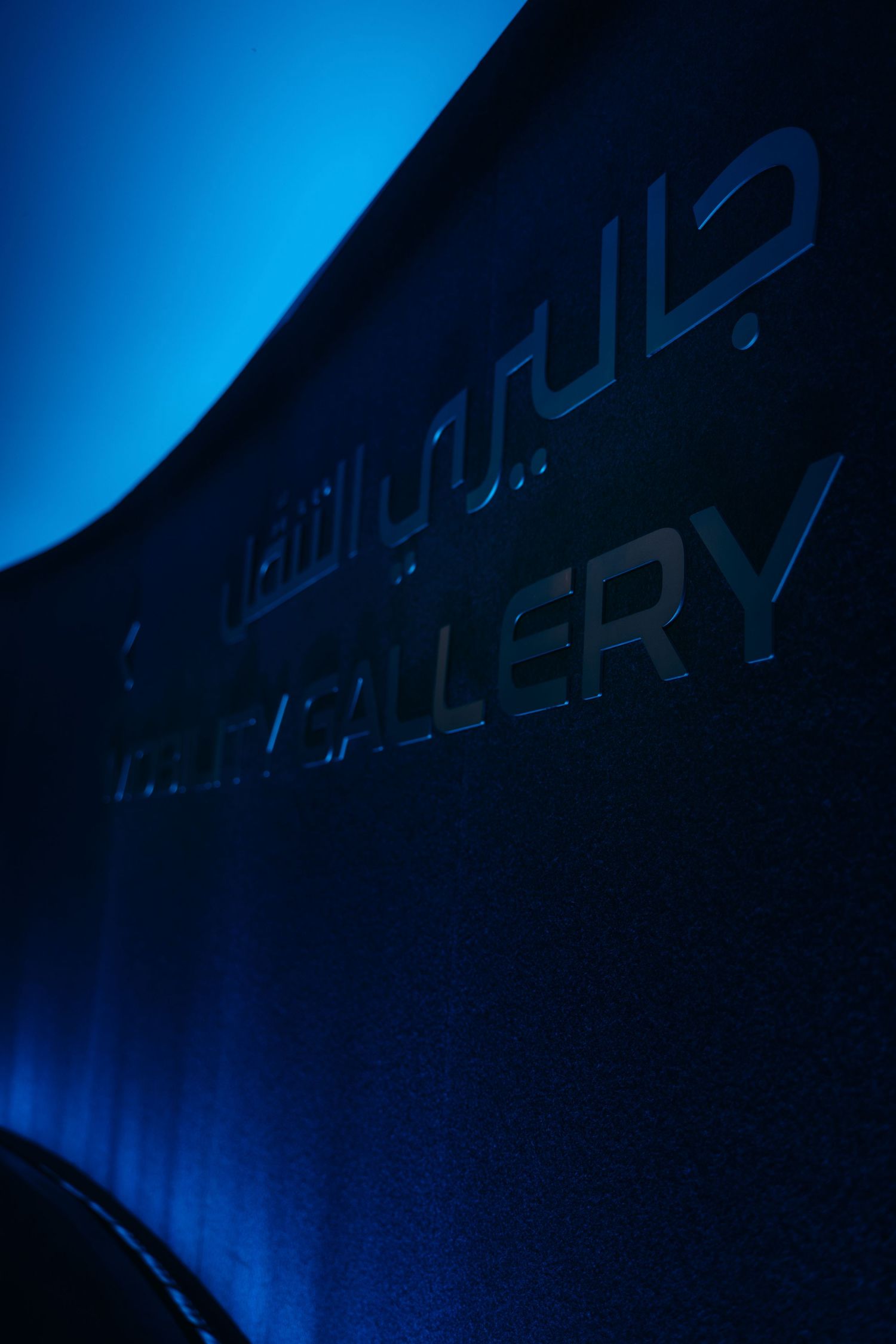 Dark blue illuminated gallery entrance sign in English and Arabic with dramatic blue lighting.