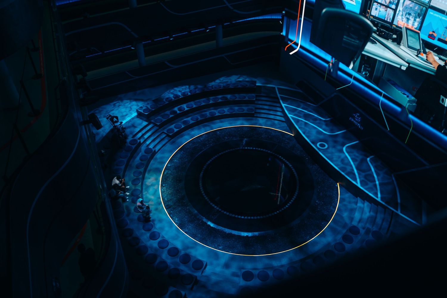 Aerial view of a futuristic circular amphitheater stage with blue neon lighting in a dark venue.