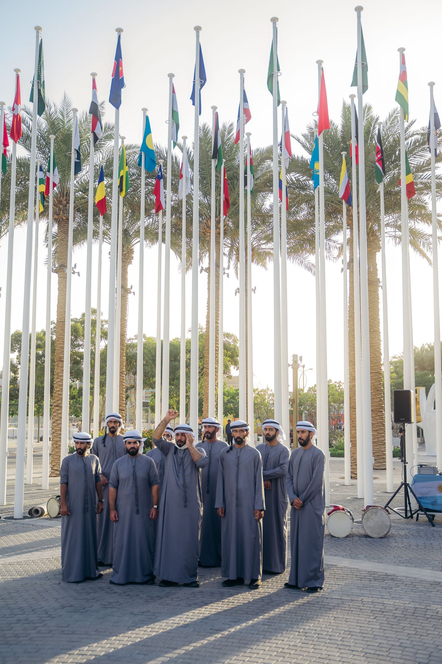 Emirati men in traditional dress stand before international flags at a cultural event in Dubai.