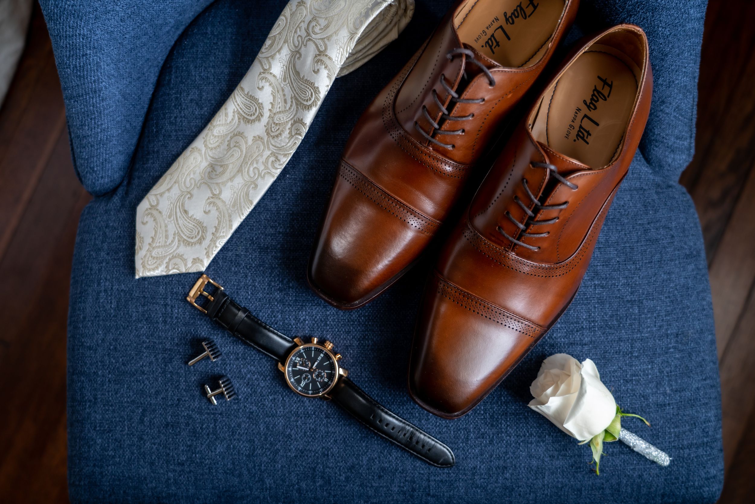 Brown leather dress shoes with tie and accessories arranged on blue fabric for a formal occasion.