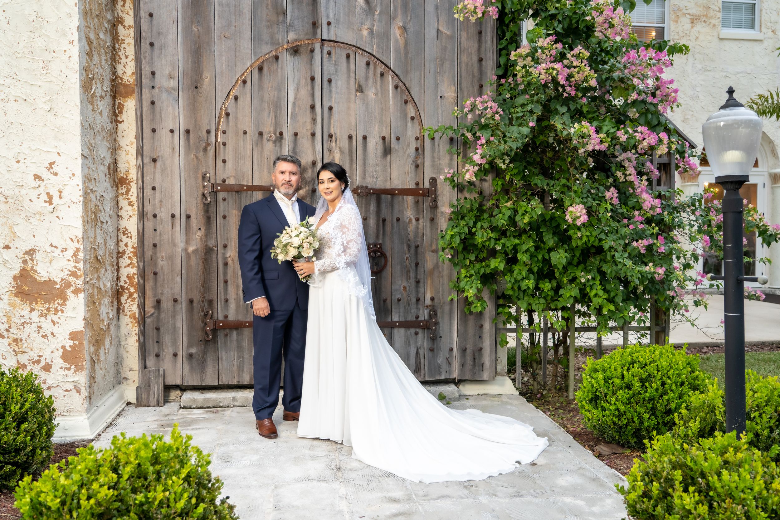 Newlyweds pose by rustic wooden door with climbing roses at historic venue.