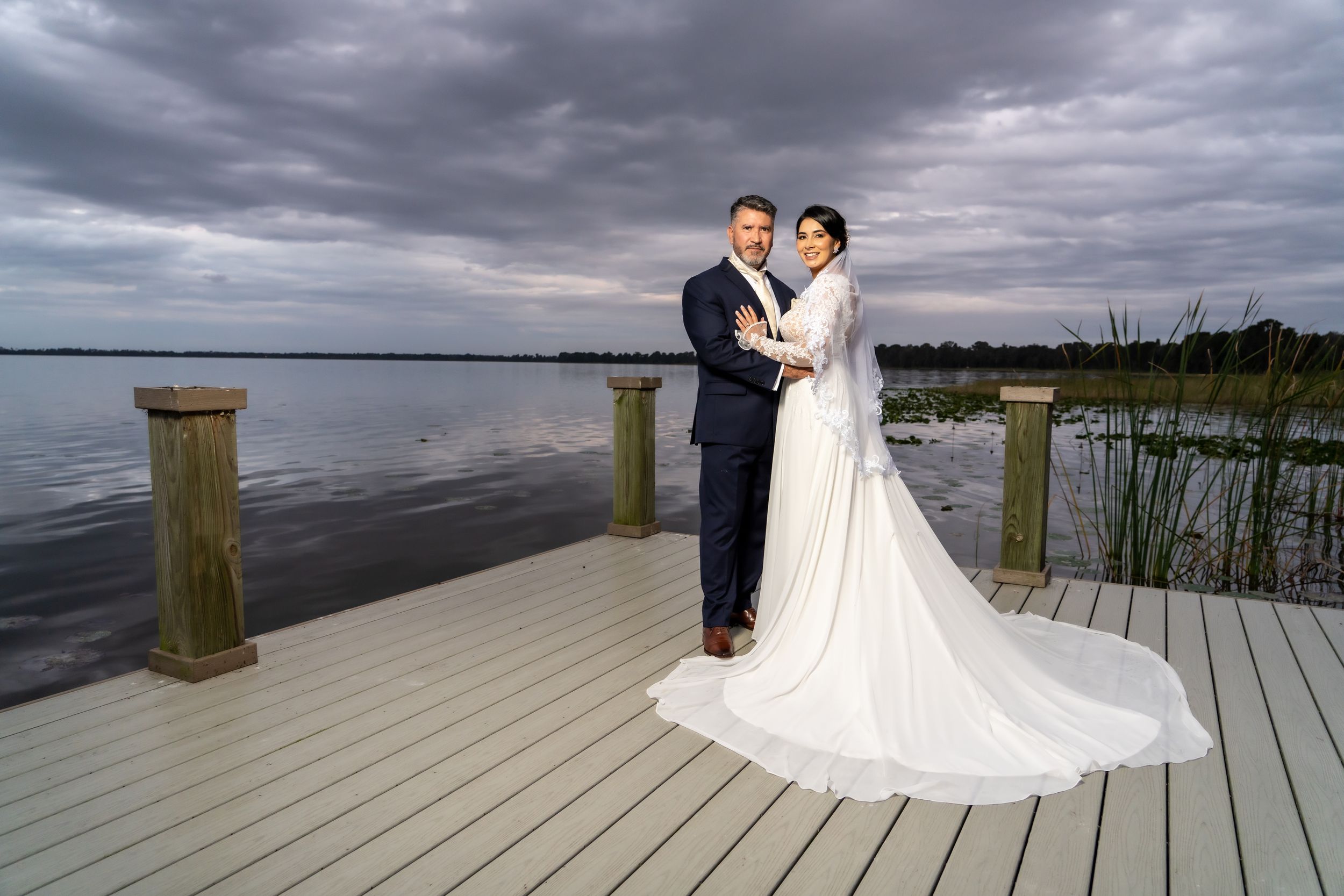 Couple in wedding attire standing on waterfront deck under dramatic cloudy sky.