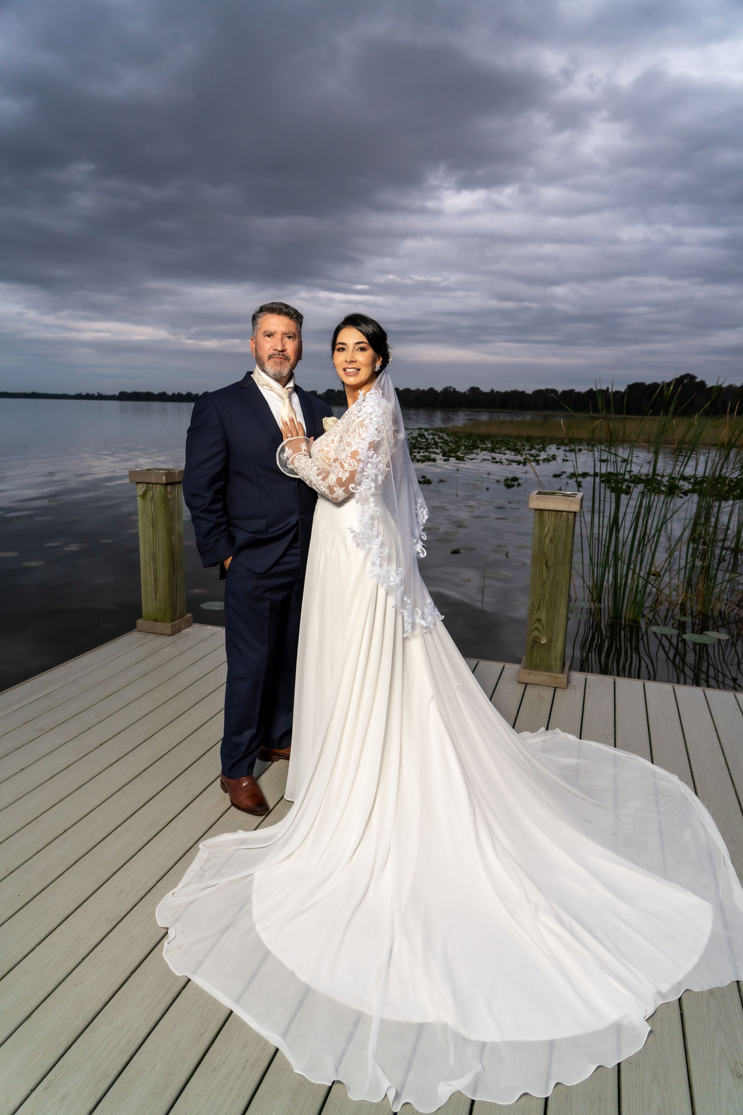 Wedding portrait on wooden dock with dramatic storm clouds and water backdrop.