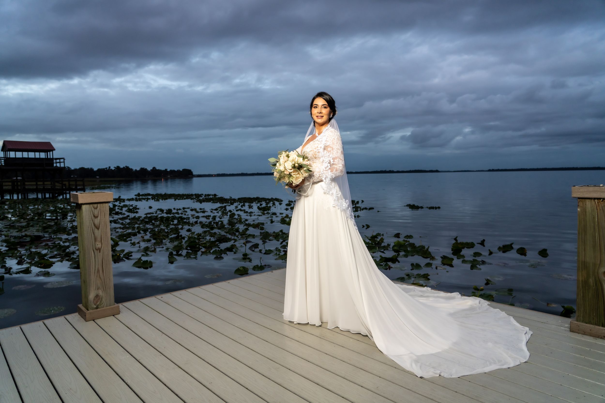 Bride in flowing white dress holds bouquet on waterfront deck under moody sky.