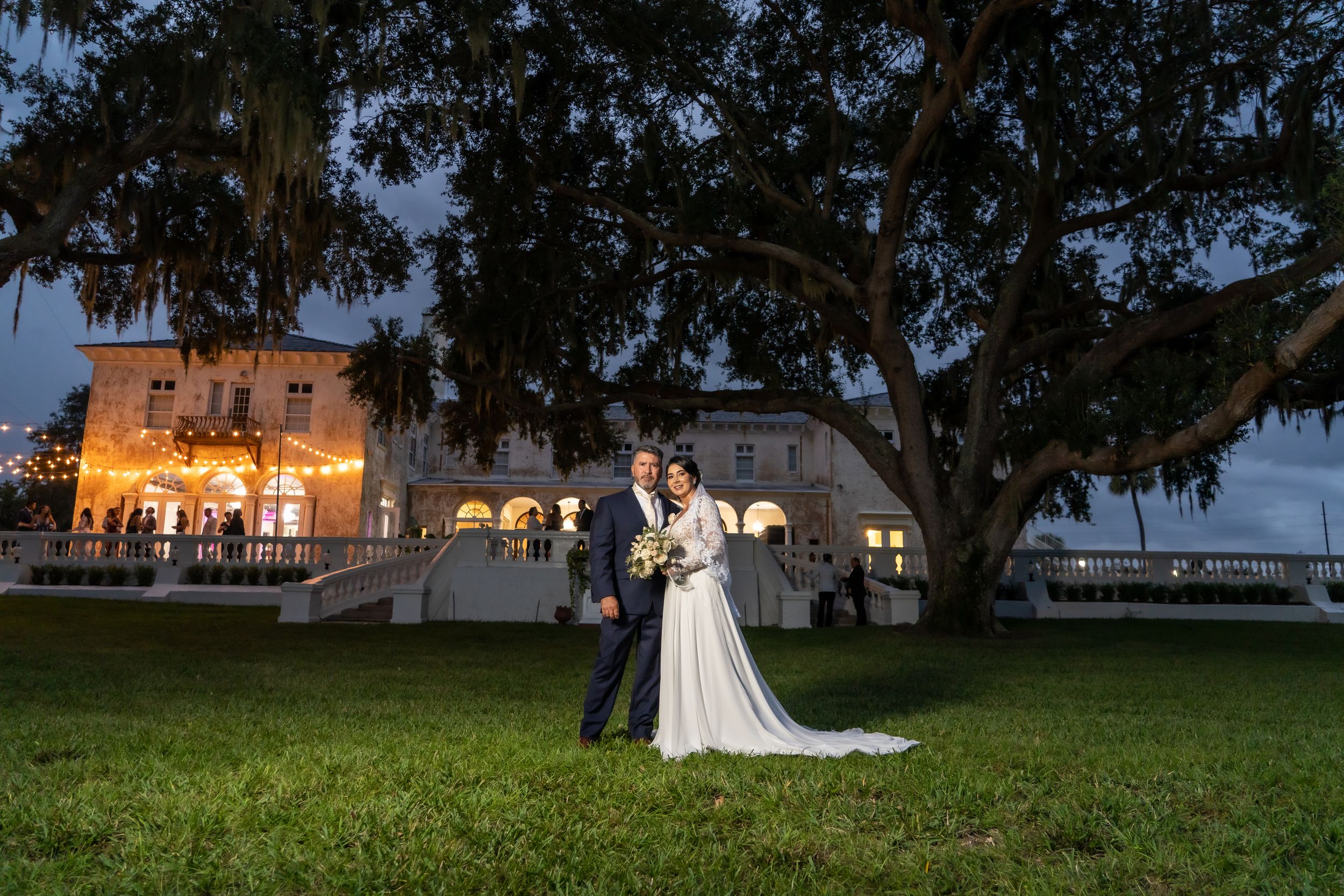 Wedding couple poses at night with illuminated historic mansion and oak tree in background.