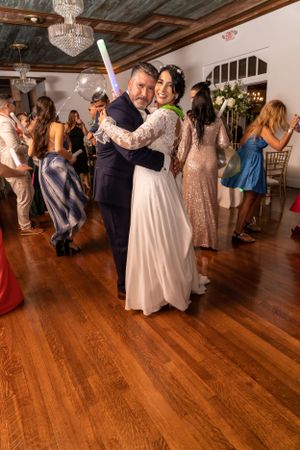 A couple dances together at a wedding reception with a chandelier overhead and guests dancing in the background.