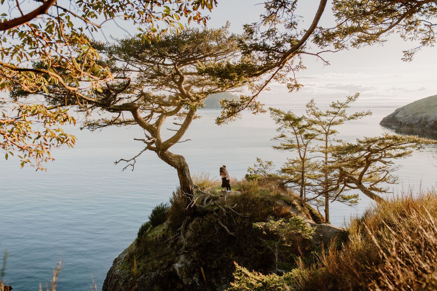 Couple standing on dramatic coastal cliff overlooking ocean bay at sunset.
