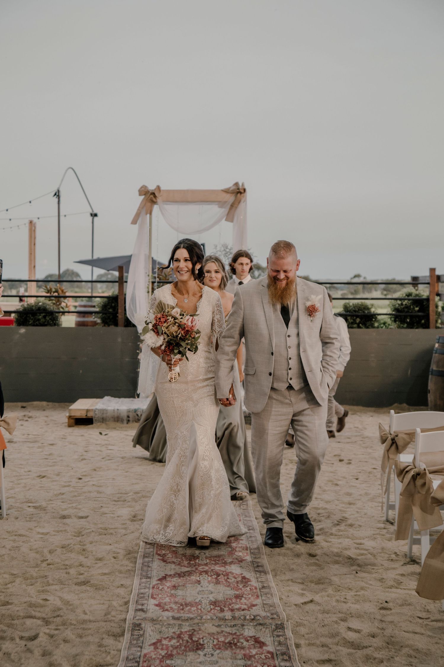 Newlywed couple walking down aisle on rooftop venue with floral arch and patterned runner at sunset ceremony.