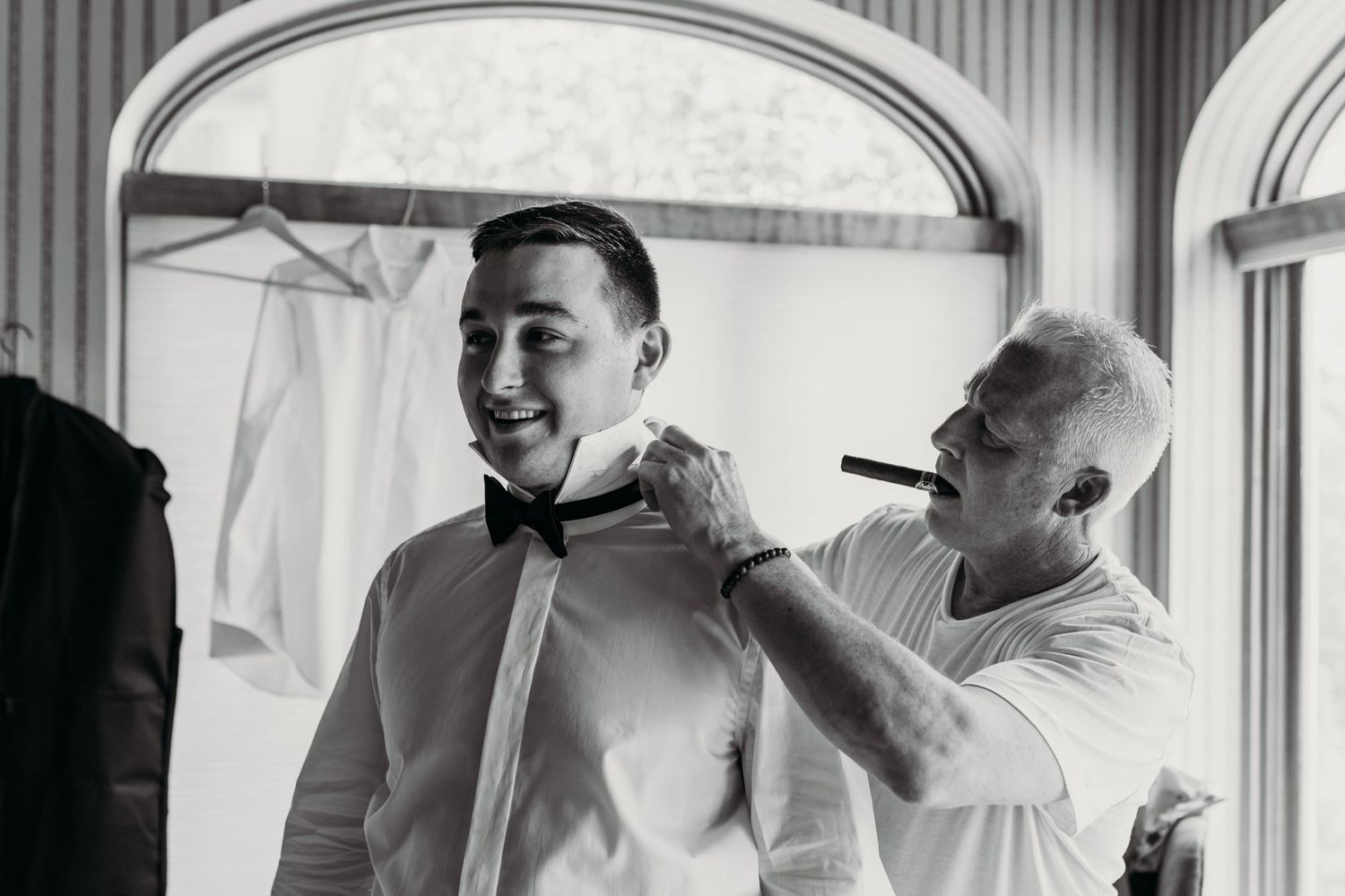 Black and white photo of two people helping with formal attire preparations indoors.