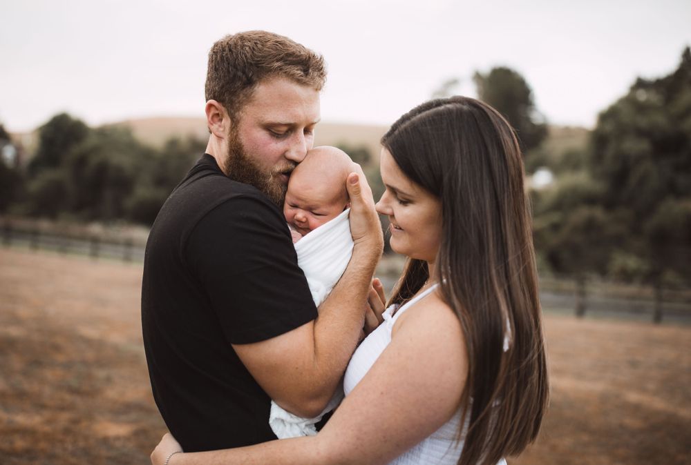 A family portrait outdoors showing parents holding their newborn baby close.