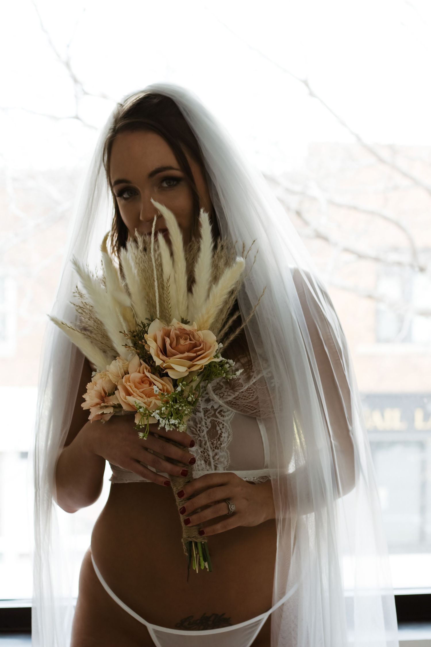 Bride in white lingerie holds romantic bouquet with pampas grass and peach roses.