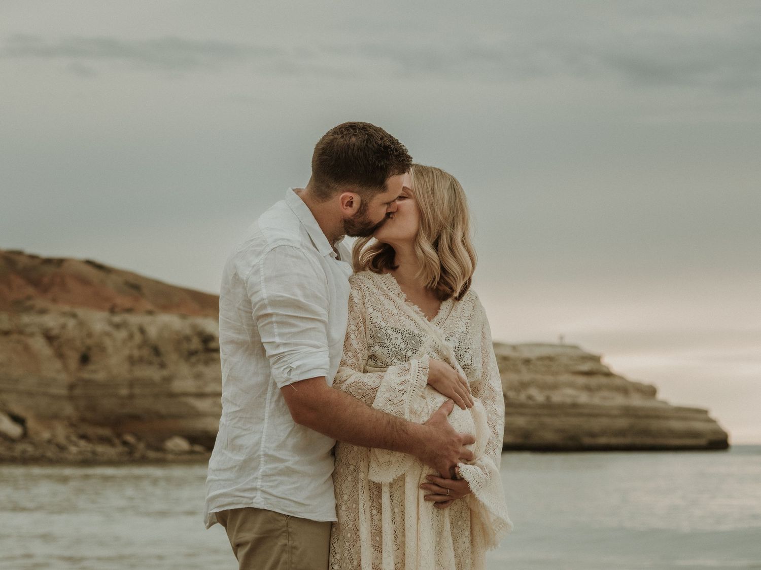 Romantic couple embracing on beach with rocky cliffs at sunset.