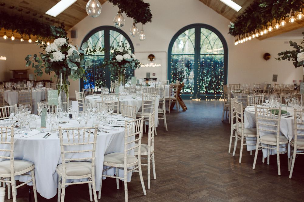 Spacious wedding reception hall with arched windows, white chairs, and crystal chandeliers.