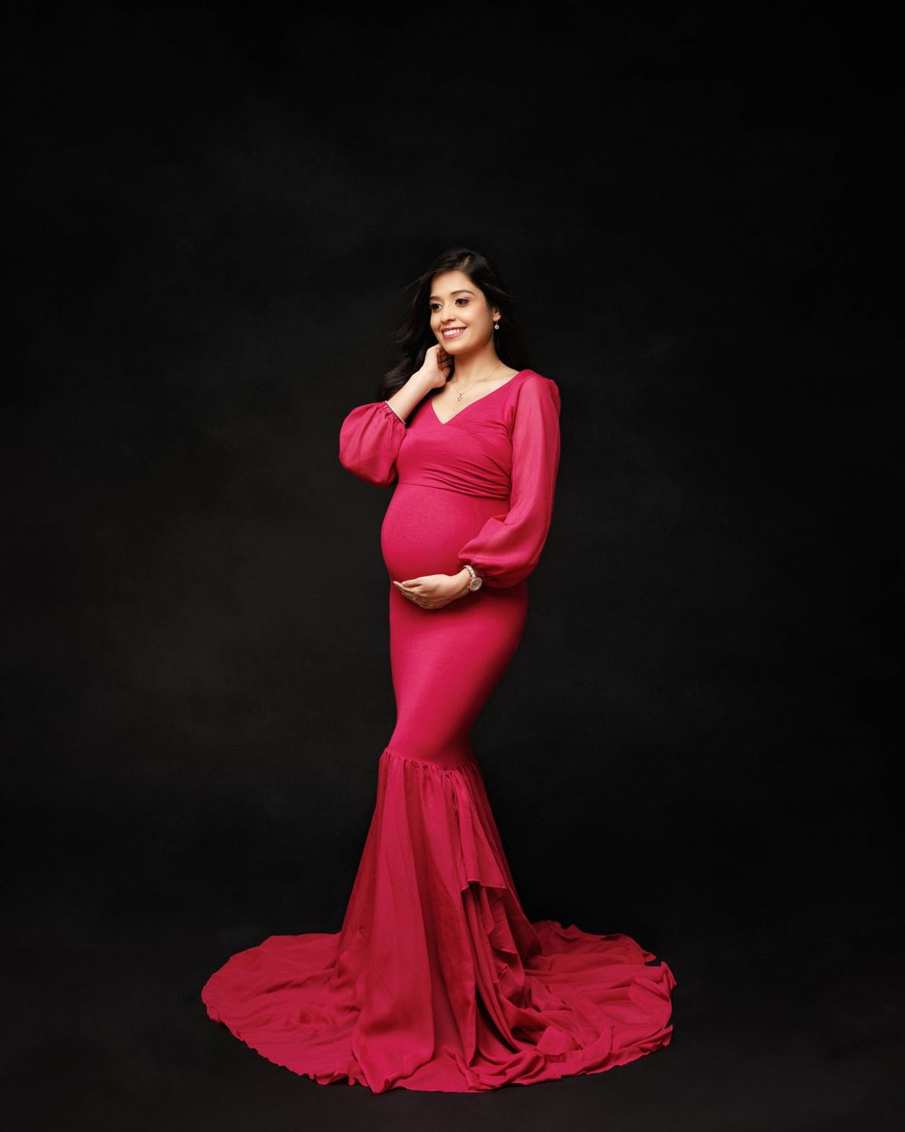 Glamorous maternity portrait in fuchsia mermaid gown with dramatic train against black backdrop.