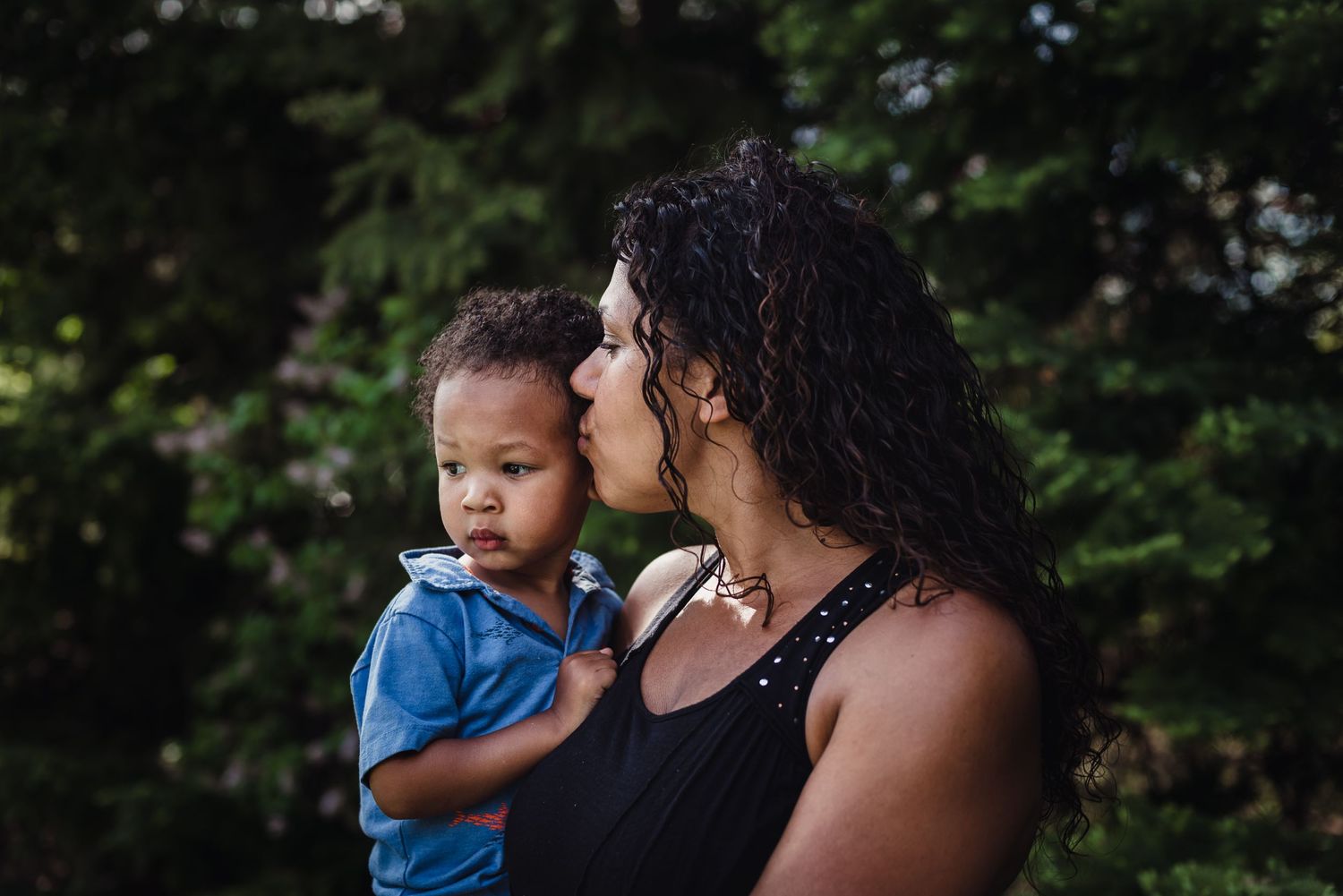 A woman kisses her young on as she holds him in her arms with pines in the background.