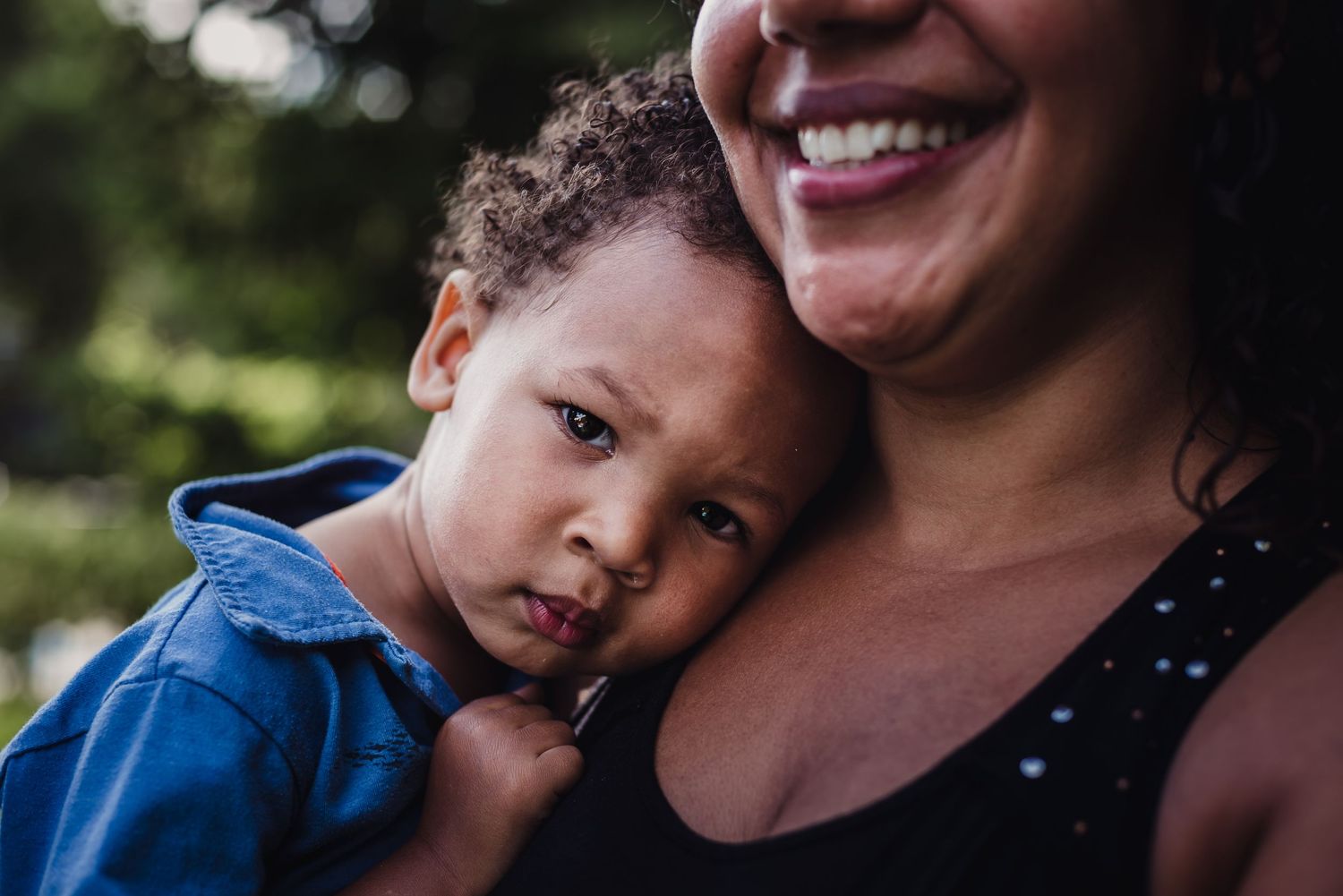 A young child leans his head on his mothers shoulder as she holds him and smiles.