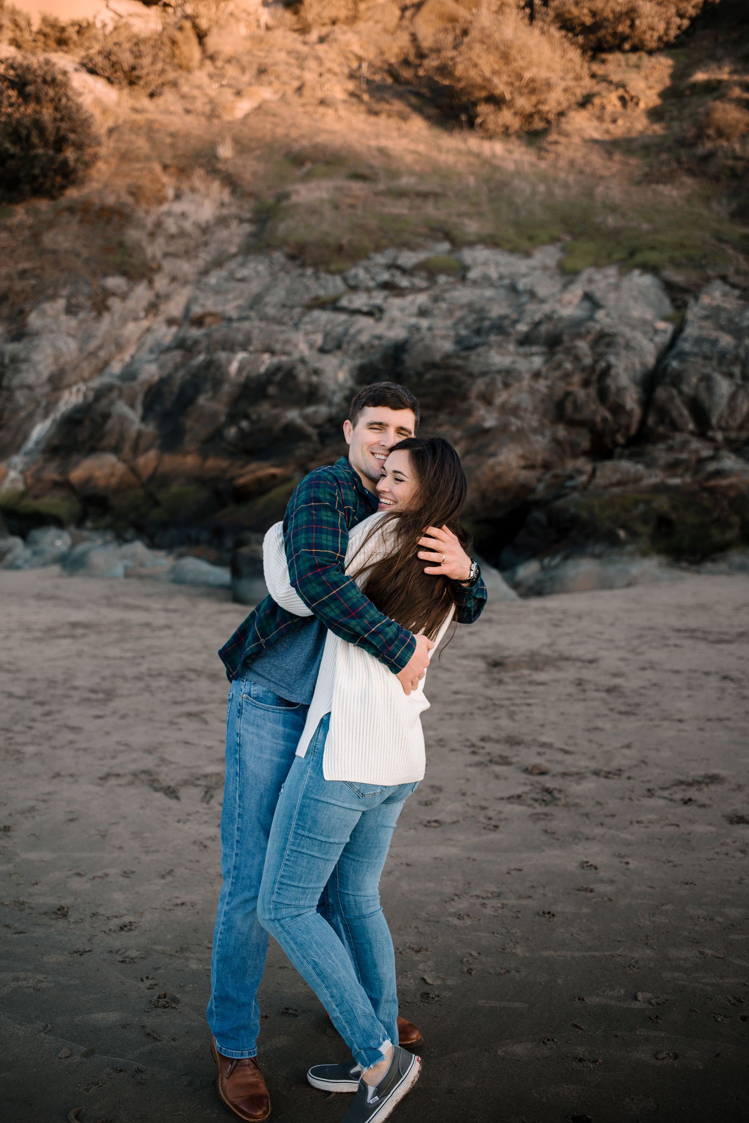 A loving embrace captured on a beach with dramatic rocky cliffs in the background.