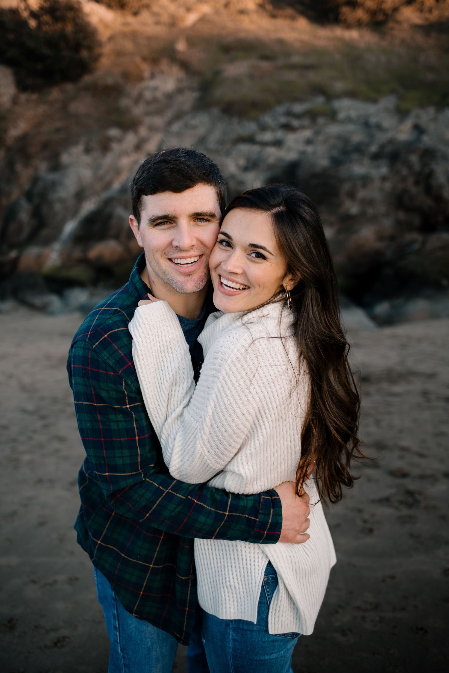 A joyful couple sharing a warm embrace on a beach with rocky cliffs during sunset.